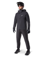 Männlicher Mountainbiker in schwarzen Alpinestars A-Dura Waterproof Pants, kombiniert mit einer passenden schwarzen wasserdichten Kapuzenjacke, schwarzem MTB-Helm mit Visier, dunkler Radsportbrille und schwarzen Handschuhen, komplette Profi-Ausrüstung für Trail-Fahrten bei jedem Wetter