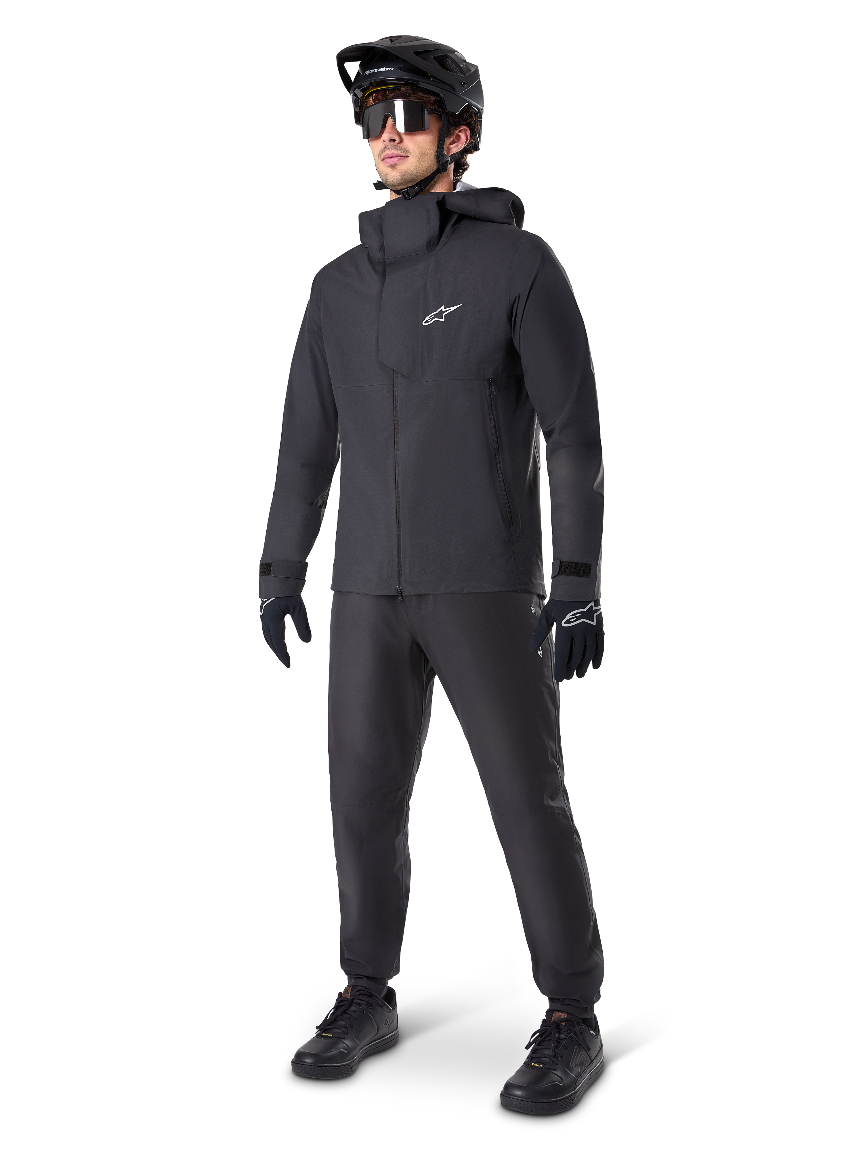 Männlicher Mountainbiker in schwarzen Alpinestars A-Dura Waterproof Pants, kombiniert mit einer passenden schwarzen wasserdichten Kapuzenjacke, schwarzem MTB-Helm mit Visier, dunkler Radsportbrille und schwarzen Handschuhen, komplette Profi-Ausrüstung für Trail-Fahrten bei jedem Wetter