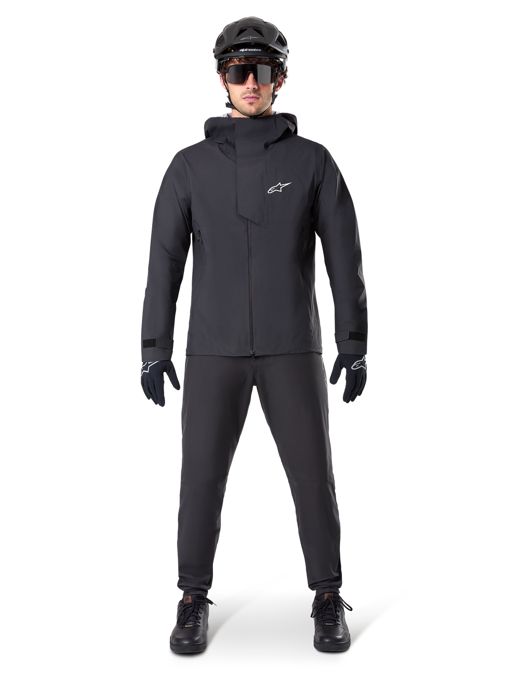 Alpinestars A-Dura wasserdichte Hose, schwarze MTB-Hose, getragen als Teil eines kompletten, ganz in Schwarz gehaltenen Mountainbike-Ausrüstungssets, einschließlich einer technischen wasserdichten Jacke, einem schwarzen Helm, einer Sonnenbrille und Handschuhen, entwickelt für Offroad-Fahrten bei nassem Wetter.