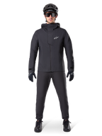 Alpinestars A-Dura wasserdichte Hose, schwarze MTB-Hose, getragen als Teil eines kompletten, ganz in Schwarz gehaltenen Mountainbike-Ausrüstungssets, einschließlich einer technischen wasserdichten Jacke, einem schwarzen Helm, einer Sonnenbrille und Handschuhen, entwickelt für Offroad-Fahrten bei nassem Wetter.