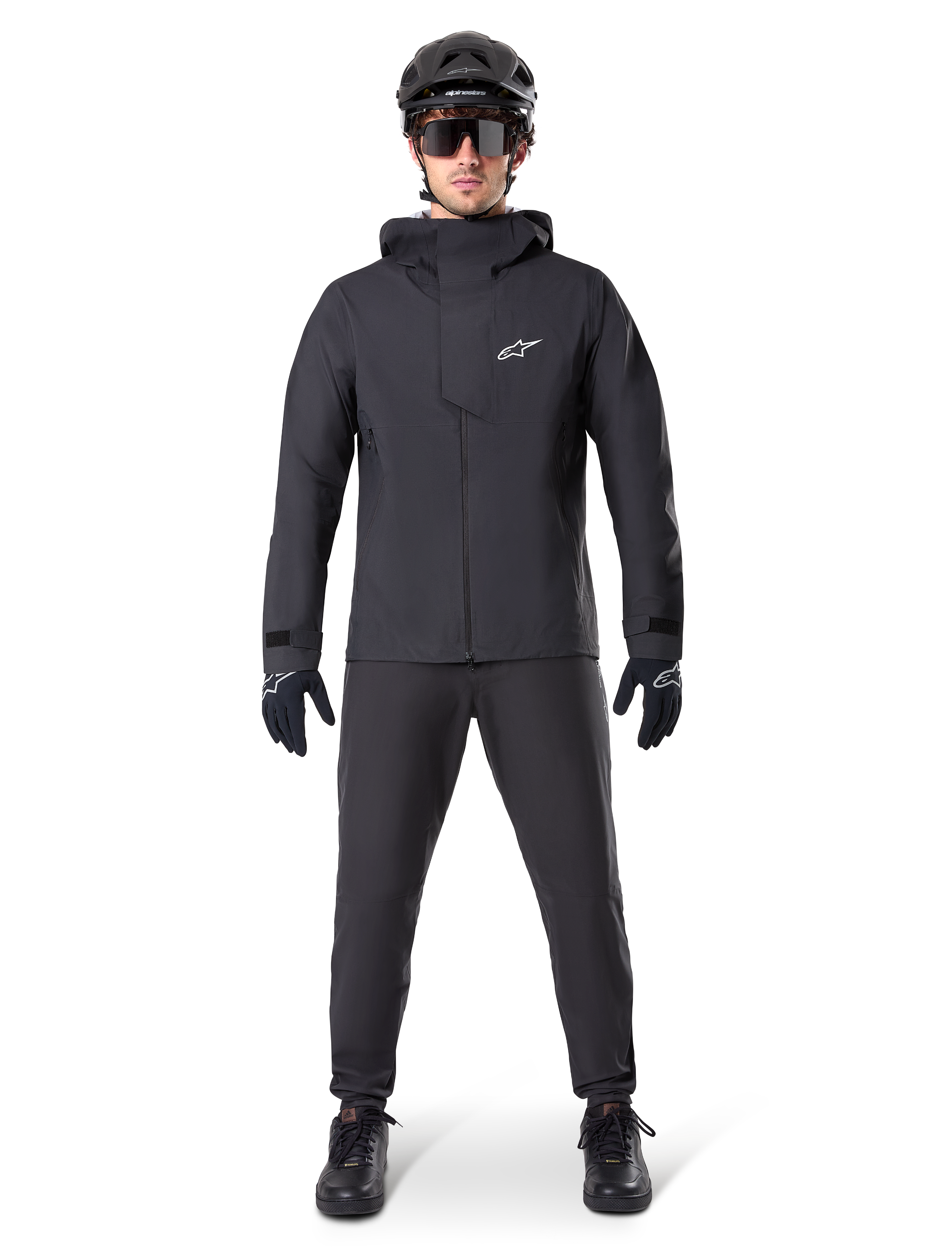 Alpinestars A-Dura wasserdichte Hose, schwarze MTB-Hose, getragen als Teil eines kompletten, ganz in Schwarz gehaltenen Mountainbike-Ausrüstungssets, einschließlich einer technischen wasserdichten Jacke, einem schwarzen Helm, einer Sonnenbrille und Handschuhen, entwickelt für Offroad-Fahrten bei nassem Wetter.
