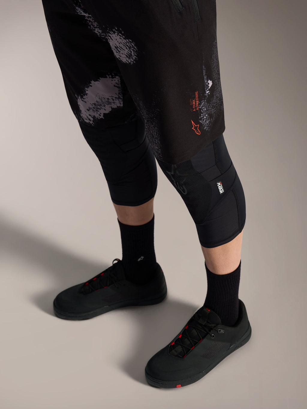 Alpinestars A-Dura Ride Shorts in Black Camo, robuste MTB-Hose mit orangefarbenem Logo, Knieschonern und Radschuhen.