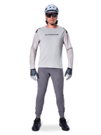 Alpinestars A-Aria Elite Hose, dunkelgraue Mountainbike-Hose, getragen von einem Fahrer in hellgrauem Langarmtrikot, hellblauem Helm und schwarzen Schuhen, mit sichtbaren Laserperforationen an den Beininnenseiten und schmaler Performance-Passform.
