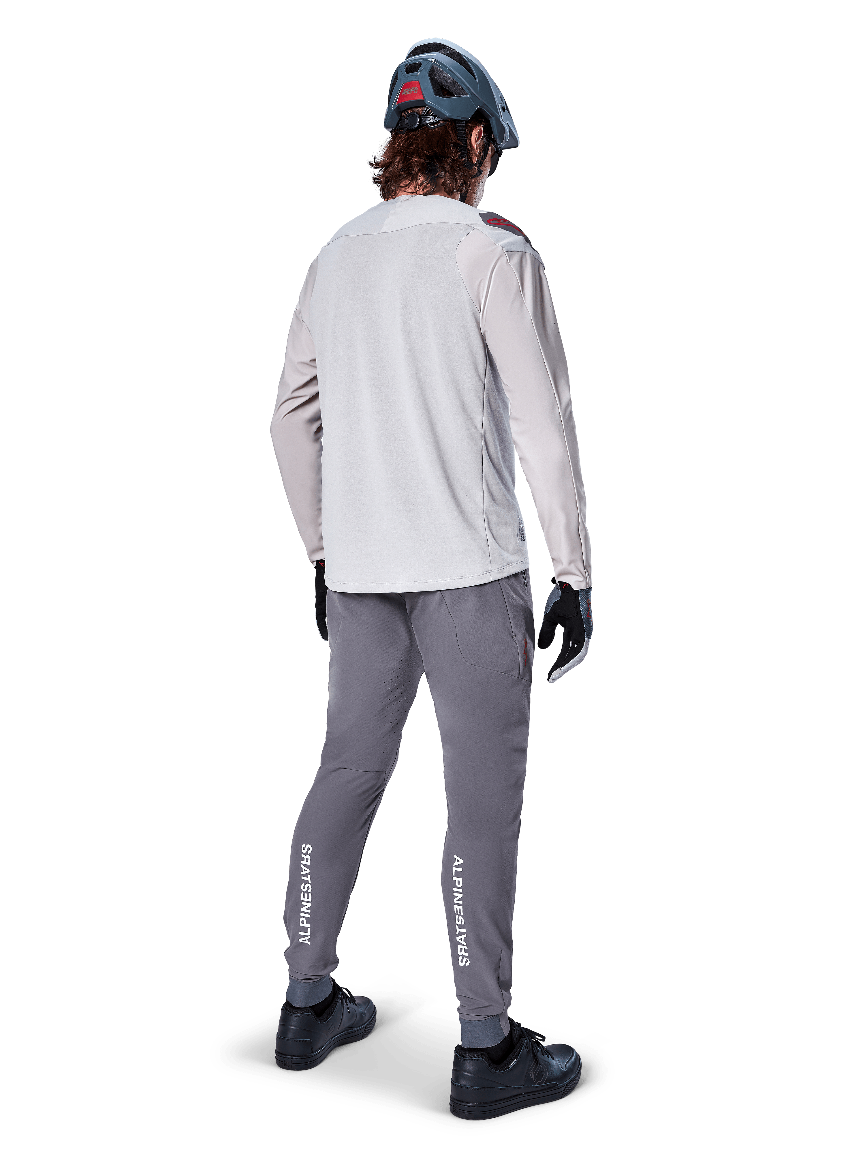 Alpinestars A-Aria Elite Hose, MTB-Fahrradhose, Dunkelgrau, Rückansicht eines Fahrers in kompletter Ausrüstung, mit weißem Alpinestars-Branding an den Unterschenkeln, Performance-Passform mit Laserperforationen, kombiniert mit einem hellgrauen Langarmtrikot und grauem MTB-Helm