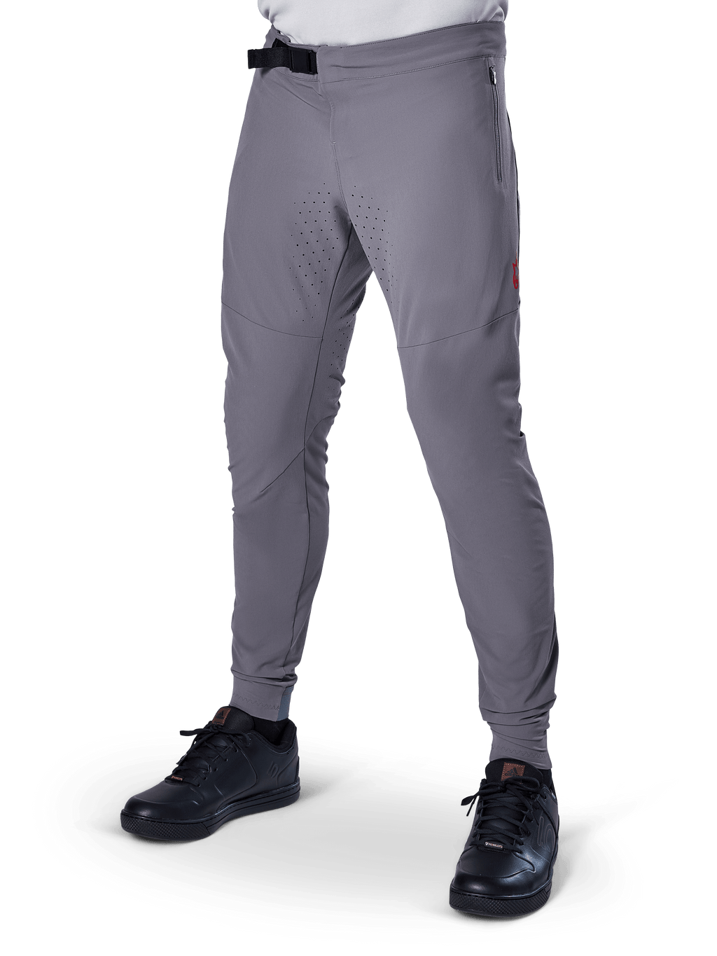 Alpinestars A-Aria Elite Hose, dunkelgraue MTB-Hose, laserperforierte Einsätze an den Oberschenkeln, schwarzer Magnetverschluss, Reißverschlusstasche, rotes Logo-Detail, schmale Performance-Passform, getragen mit schwarzen Schuhen
