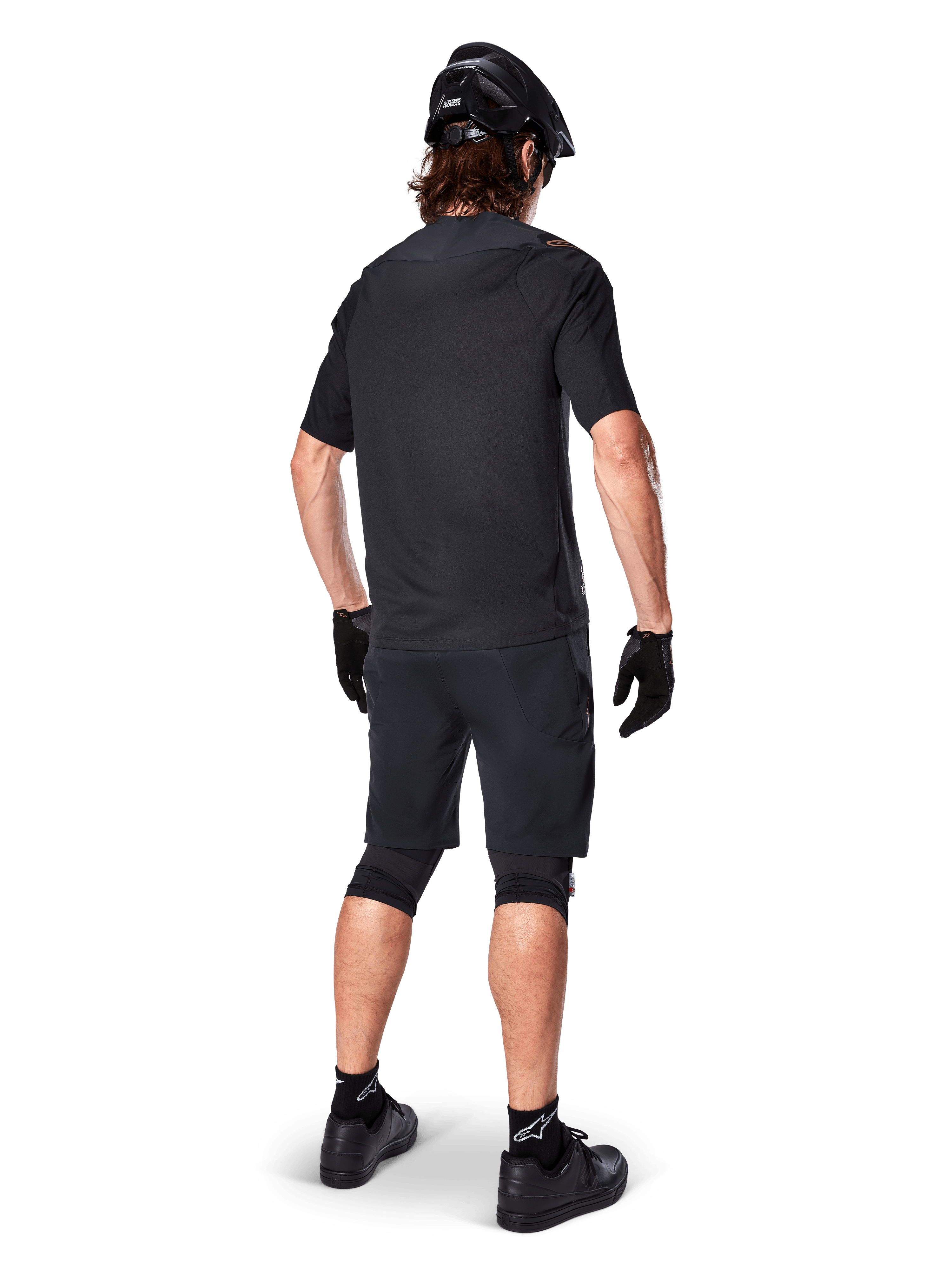 Rear view of a mountain biker wearing Alpinestars A-Aria Elite Shorts in Black, paired with a matching black short-sleeve jersey, black helmet, gloves, and MTB shoes, showing a complete performance outfit for off-road cycling.