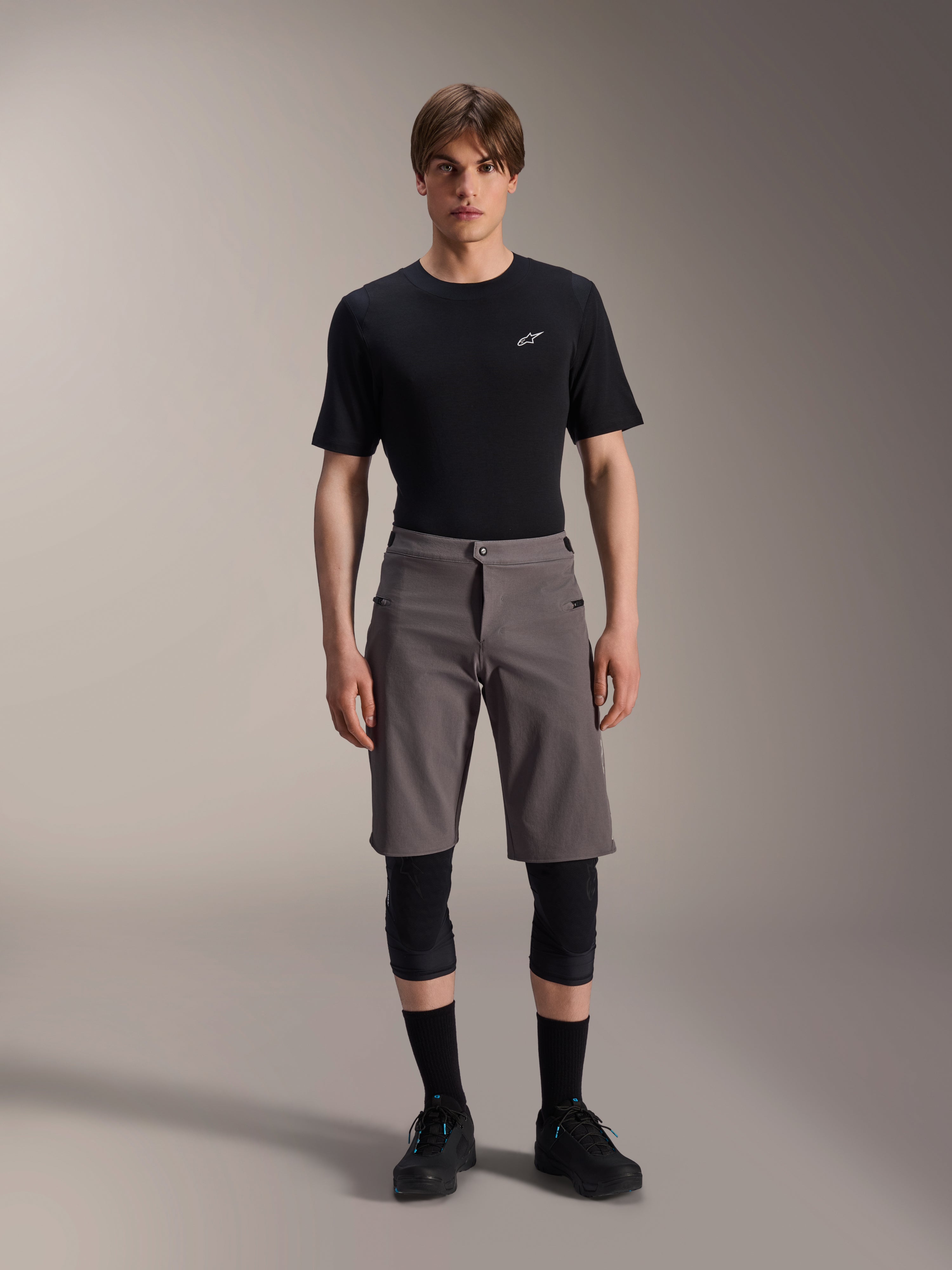 Alpinestars A-Dura Elite Shorts, dunkelgraue Mountainbike-Shorts, getragen mit einem schwarzen T-Shirt mit Rundhalsausschnitt und schwarzen Knieschützern, knielange Performance-Radsportbekleidung mit Druckknopfverschluss vorne und strapazierfähigem Cordura-Gewebe.