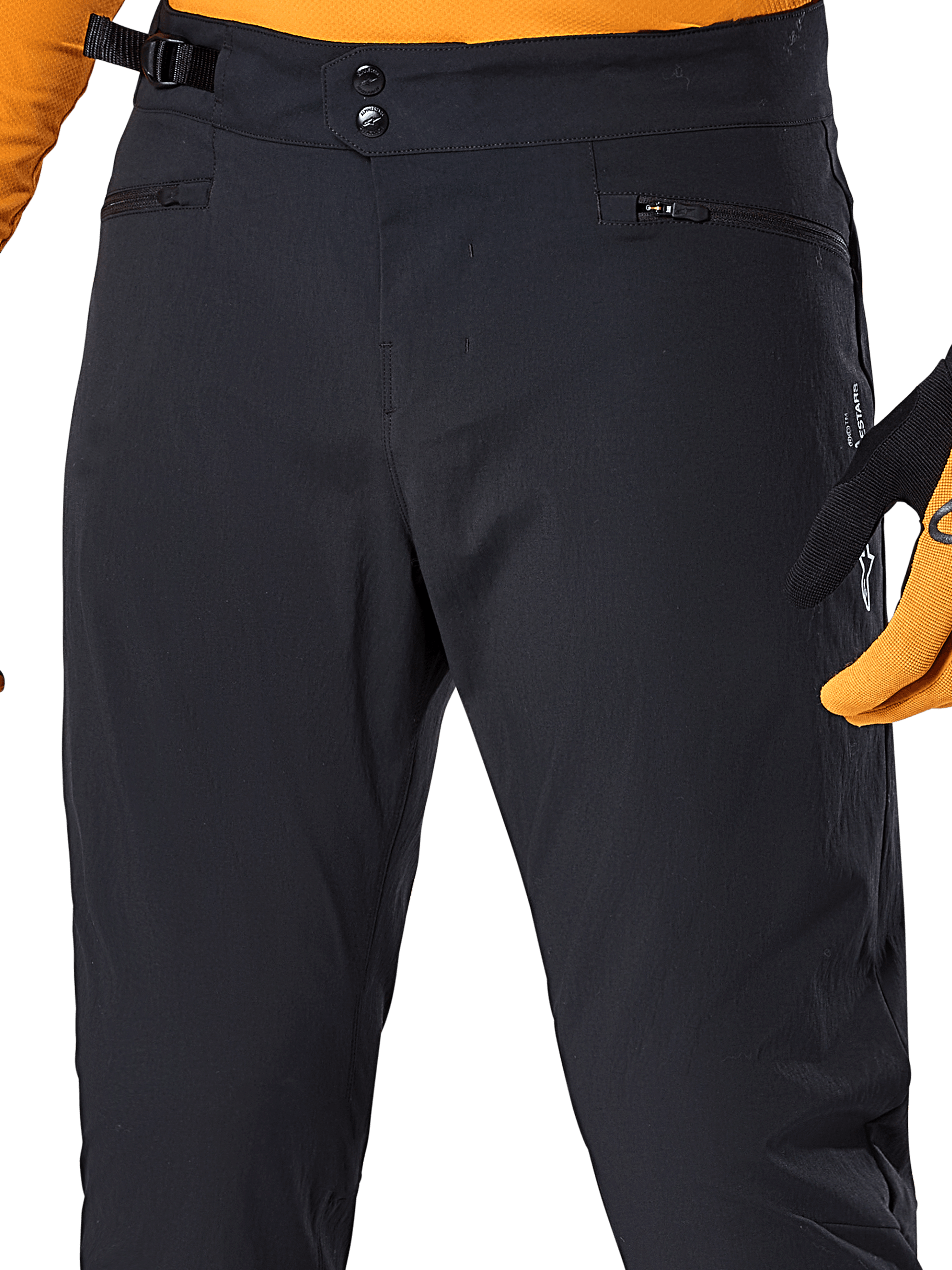 Alpinestars A-Dura Hose, schwarze MTB-Radhose, Detailansicht mit doppeltem Druckknopfverschluss, verstellbarem seitlichem Taillenversteller und Reißverschlusstaschen, kombiniert mit einem orangefarbenen Trikot und Handschuhen