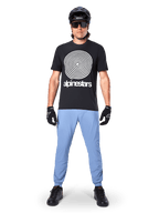 Alpinestars A-Dura Hose, Infinity Blue, MTB-Hose getragen mit einem schwarzen T-Shirt mit Spiralgrafik, schwarzen Handschuhen und einem schwarzen Helm, aus strapazierfähigem Stretch-Gewebe und mit elastischen Bündchen, Performance-Mountainbike-Ausrüstung