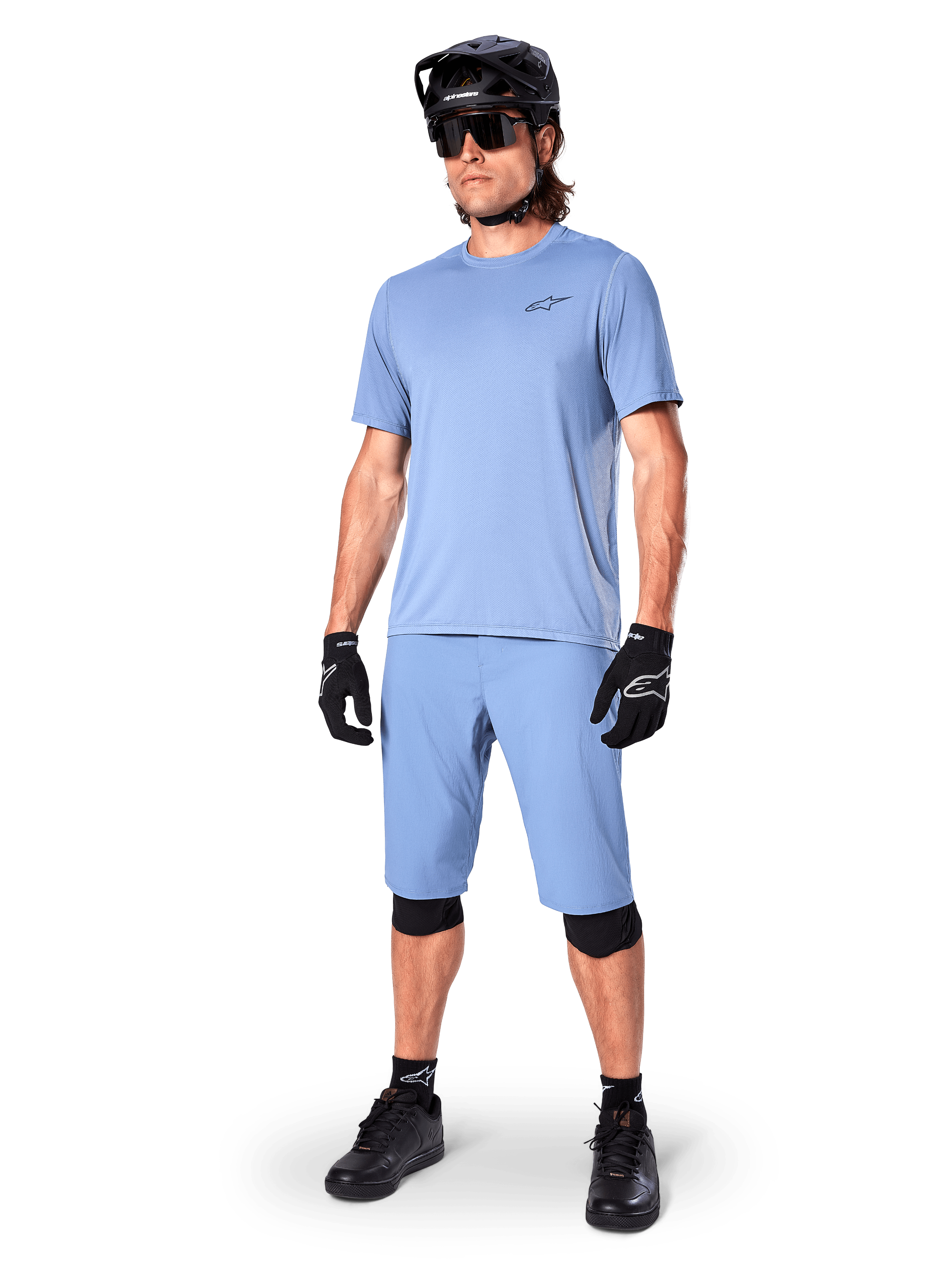 Model in Alpinestars A-Dura Shorts in Infinity Blue, passendes hellblaues Kurzarm-Trikot, schwarzer MTB-Helm, Sonnenbrille und schwarze Handschuhe, komplettes Mountainbike-Outfit für Trailfahrten