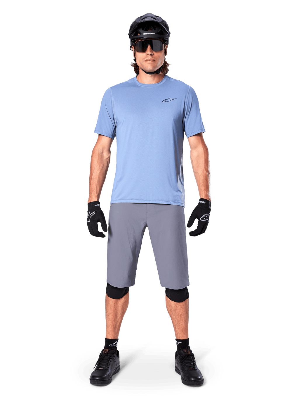 Männlicher Fahrer in Alpinestars A-Dura Shorts in Dunkelgrau, kombiniert mit einem hellblauen Kurzarm-T-Shirt, schwarzem Helm, Sonnenbrille und schwarzen Knieschützern, komplettes Mountainbike-Outfit für Offroad-Performance.
