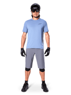 Männlicher Fahrer in Alpinestars A-Dura Shorts in Dunkelgrau, kombiniert mit einem hellblauen Kurzarm-T-Shirt, schwarzem Helm, Sonnenbrille und schwarzen Knieschützern, komplettes Mountainbike-Outfit für Offroad-Performance.