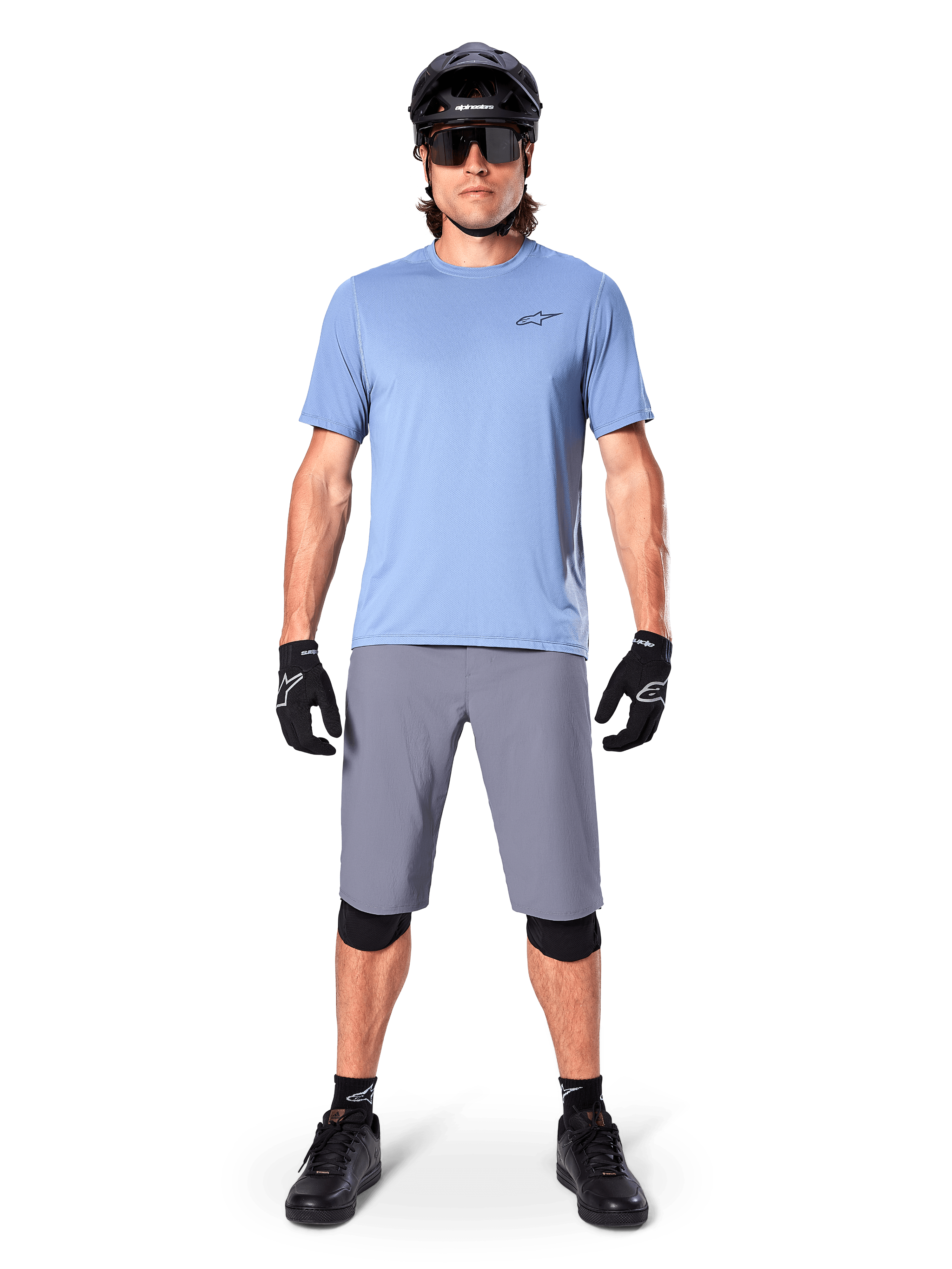 Männlicher Fahrer in Alpinestars A-Dura Shorts in Dunkelgrau, kombiniert mit einem hellblauen Kurzarm-T-Shirt, schwarzem Helm, Sonnenbrille und schwarzen Knieschützern, komplettes Mountainbike-Outfit für Offroad-Performance.