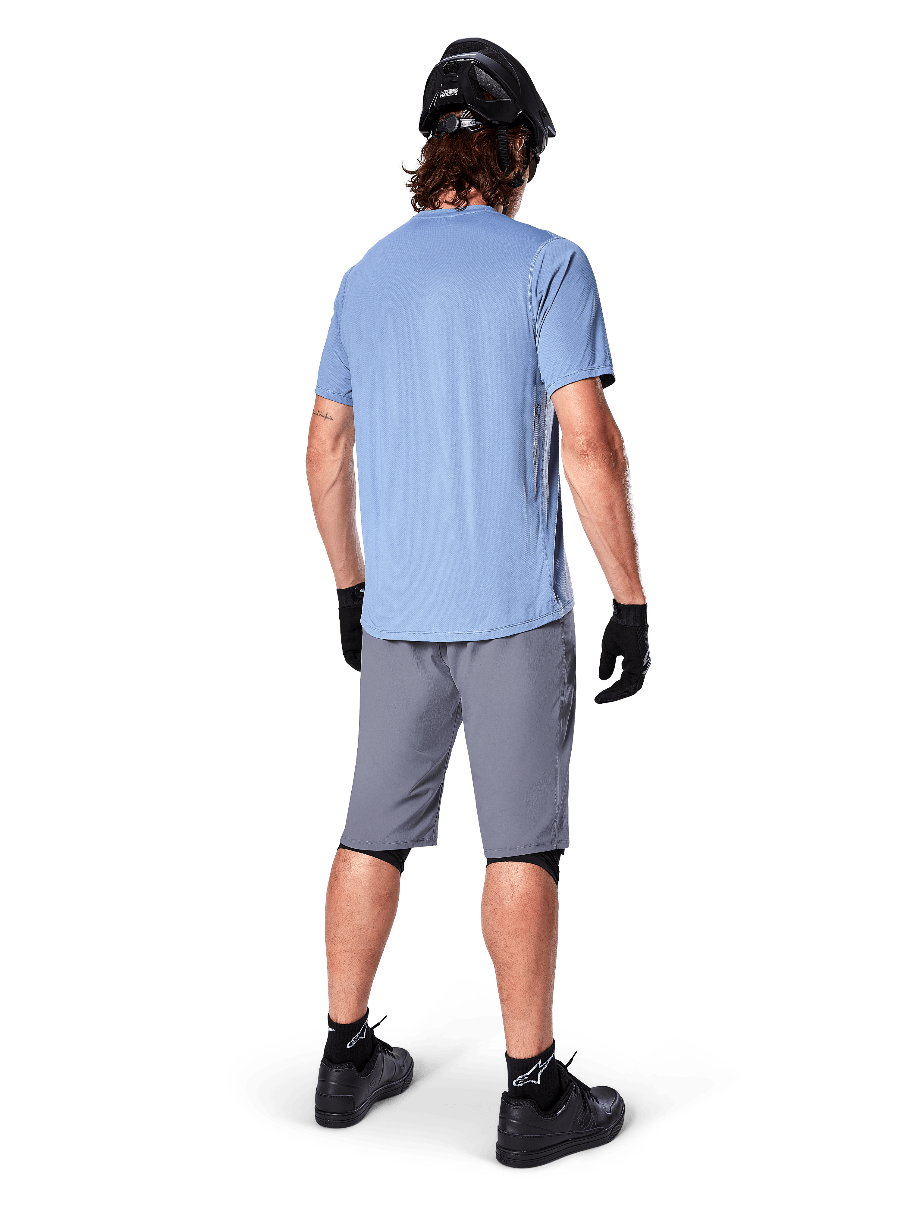 Alpinestars A-Dura Shorts, dunkelgraue MTB-Shorts, Rückansicht, getragen mit einem hellblauen Kurzarmtrikot, schwarzem Mountainbike-Helm, schwarzen Handschuhen, Socken und Schuhen, funktionelle und strapazierfähige Ausrüstung für den Offroad-Radsport