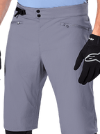 Alpinestars A-Dura Shorts, MTB-Shorts, Dunkelgrau, Nahaufnahme der Taille mit doppeltem Druckknopfverschluss, seitlichem Taillenversteller und Reißverschlusstaschen, kombiniert mit schwarzen Fahrradhandschuhen, strapazierfähige Mountainbike-Bekleidung.