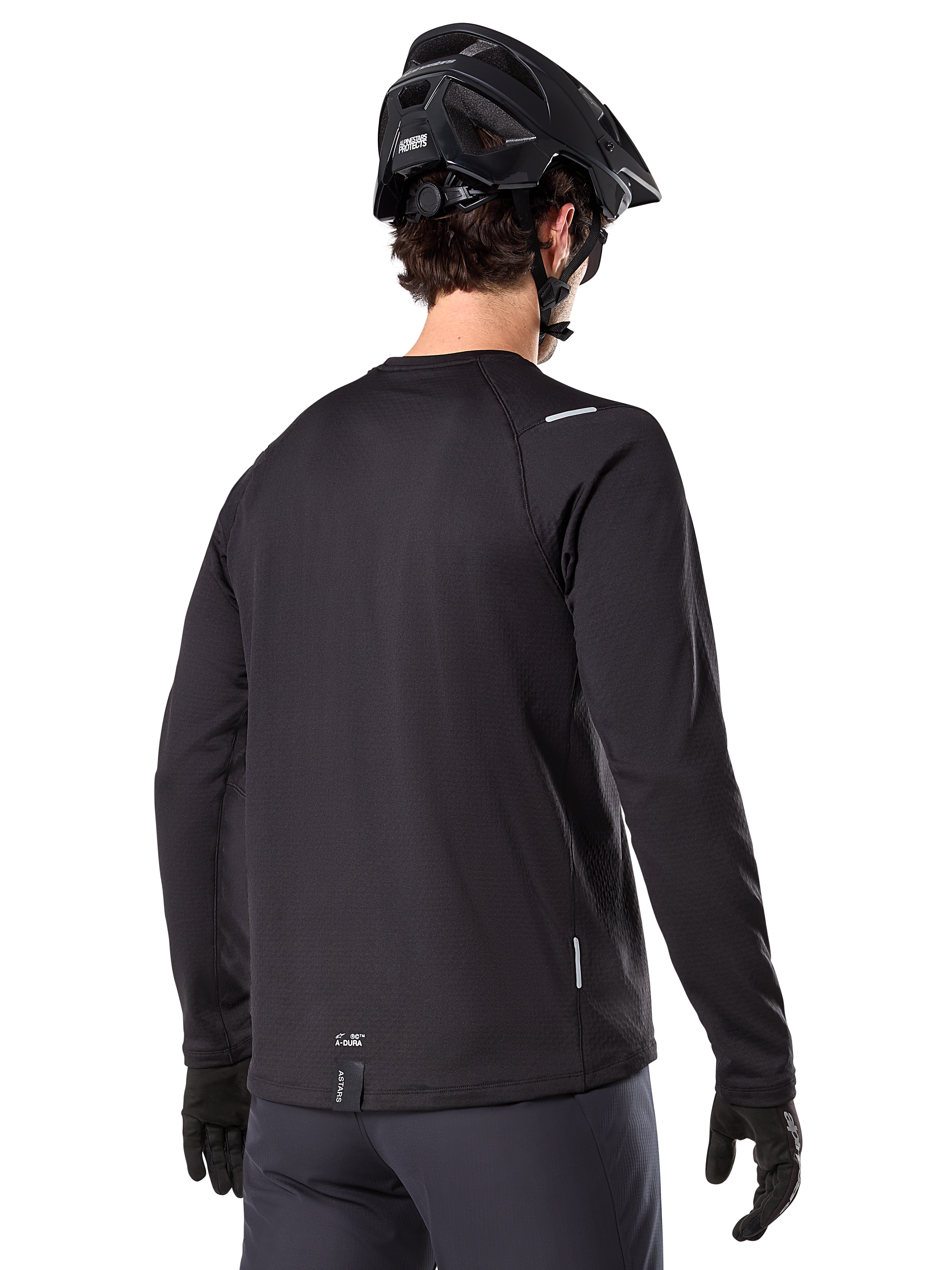 Alpinestars A-Dura Thermal Jersey, langärmeliges Mountainbike-Trikot, Schwarz, Rückansicht mit verlängertem Rückenteil, kleine reflektierende Details und Branding am Saum, getragen mit einem schwarzen Helm und Handschuhen für das Radfahren bei kaltem Wetter