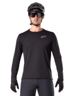 Alpinestars A-Dura Thermal Trikot, schwarzes langärmeliges Mountainbike-Trikot, getragen mit einem schwarzen Helm und einer großen Sonnenbrille, mit kleinem weißen Alpinestars-Logo auf der Brust, reguläre Passform, MTB-Radsportbekleidung