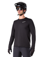 Alpinestars A-Dura Thermal Jersey, schwarzes Langarm-Mountainbike-Trikot, weißes Brust-Logo, getragen mit schwarzem Helm und Handschuhen, schlichtes sportliches Design für Trail-Fahrten