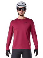 Männlicher Radfahrer trägt das Alpinestars A-Dura Thermal Jersey, langärmeliges MTB-Trikot, Burgunderrot, kombiniert mit einem grauen Helm und einer schwarzen Sonnenbrille, kleines schwarzes Stern-Logo auf der Brust, isoliertes Fleece-Material für Mountainbiking bei kaltem Wetter