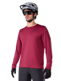 Alpinestars A-Dura Thermal Jersey, Langarm-Mountainbike-Trikot, Burgunderrot, mit isoliertem Fleece-Futter und kleinem schwarzem Brust-Logo, getragen mit einem grauen Helm, schwarzer Sonnenbrille und schwarzen Handschuhen, ideal für Radsport bei kaltem Wetter