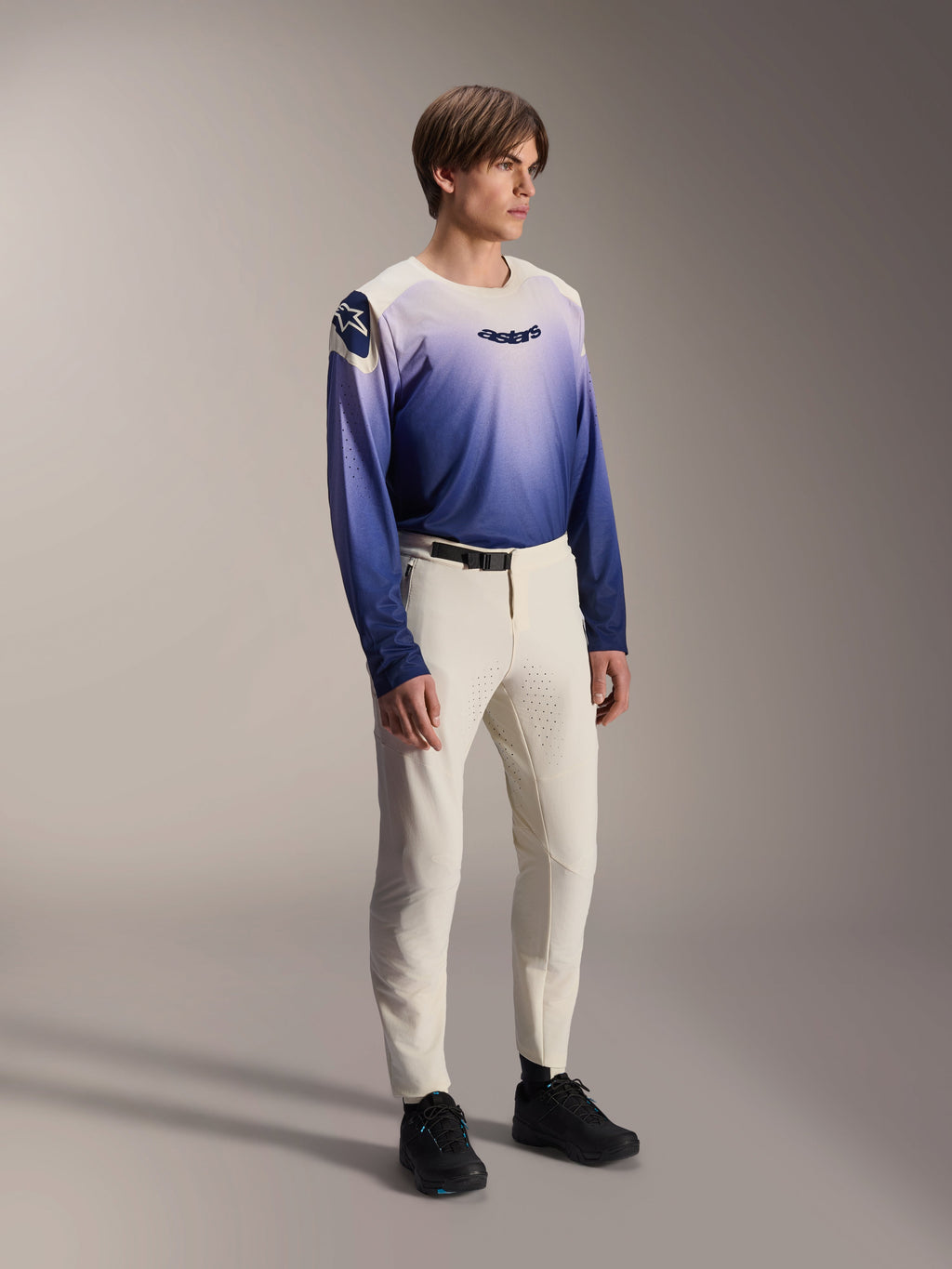 Alpinestars A-Aria Polartec Maker Elite Jersey in Baja Blue, männliches Model, Design mit Farbverlauf von Weiß zu Blau und schwarzem 'astars'-Logo auf der Brust, kombiniert mit cremeweißen Mountainbike-Hosen und schwarzen Schuhen, Performance-Passform MTB-Bekleidung für Trail-Fahrten.