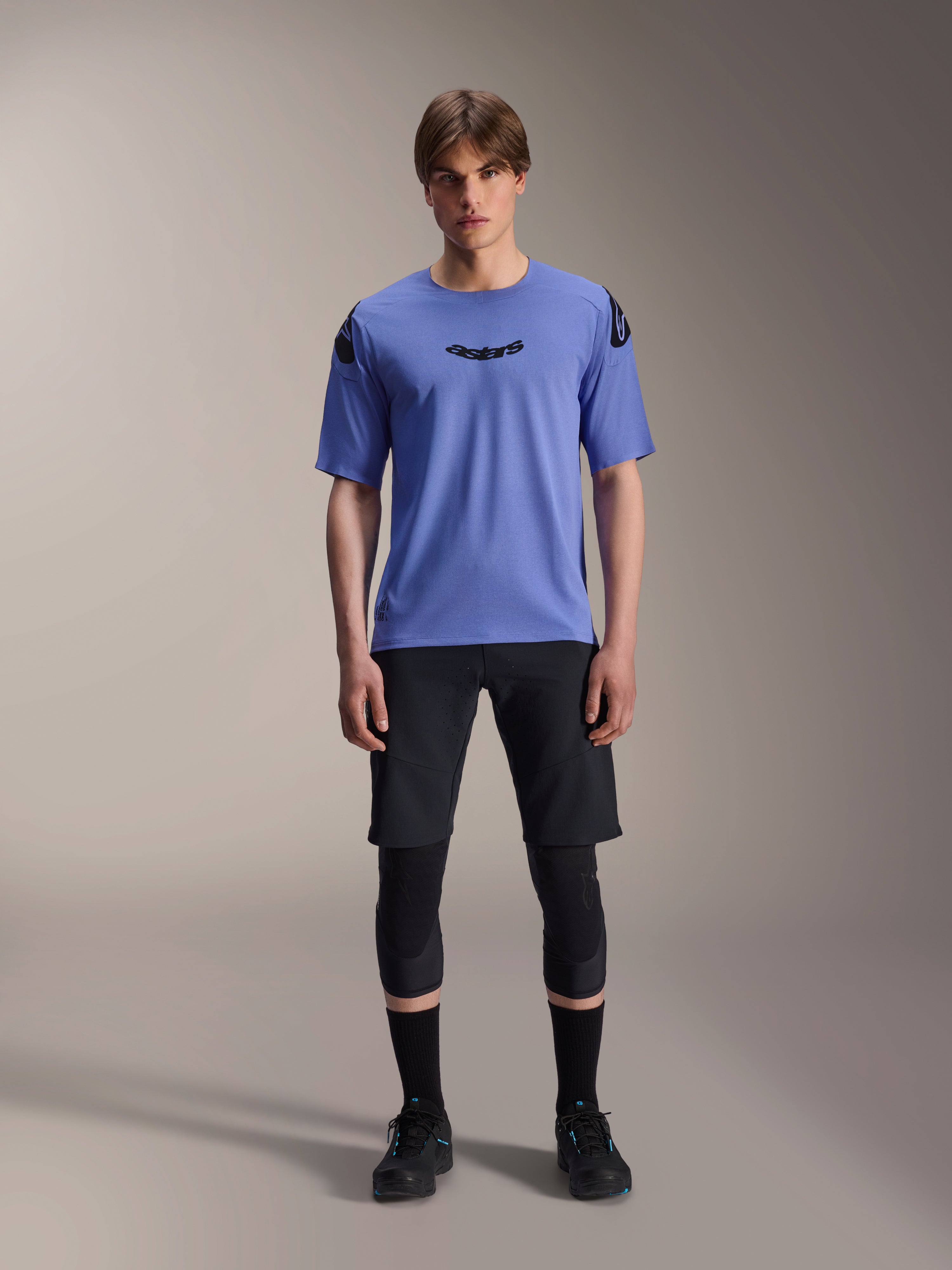 Alpinestars A-Aria Elite Shorts, MTB Mountainbike-Shorts, Black Titanium, getragen mit blauem Kurzarmtrikot, Knieschützern und schwarzen Bikeschuhen, Ganzkörper-Studioaufnahme, Performance-Mountainbike-Bekleidung