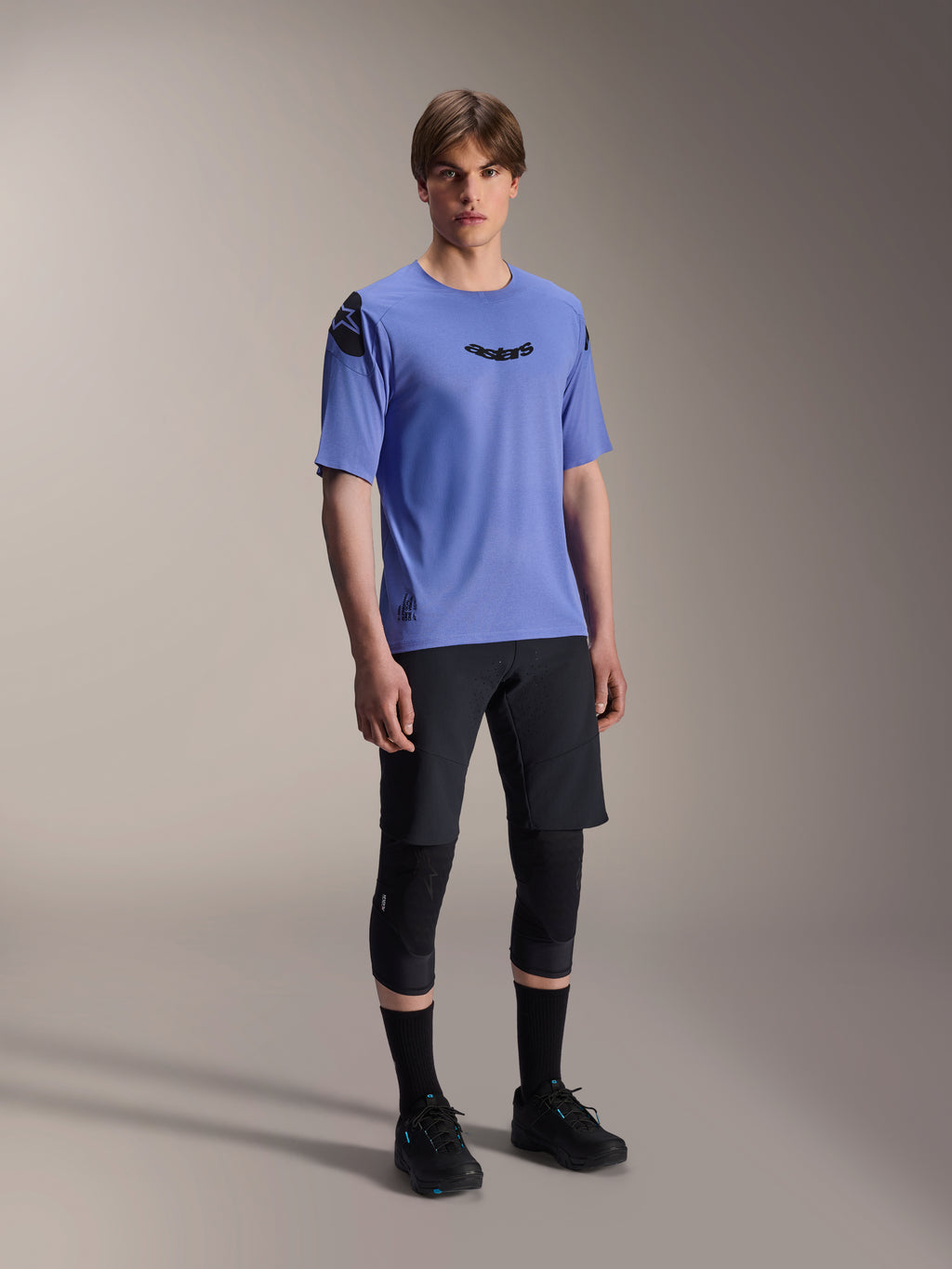Männliches Modell in Alpinestars A-Aria Elite Shorts in Black Titanium und A-Aria Polartec Elite SS Jersey in Helllila, kombiniert mit schwarzen Mountainbike-Knieschützern und Schuhen, professionelle Radsportausrüstung für Offroad-Performance