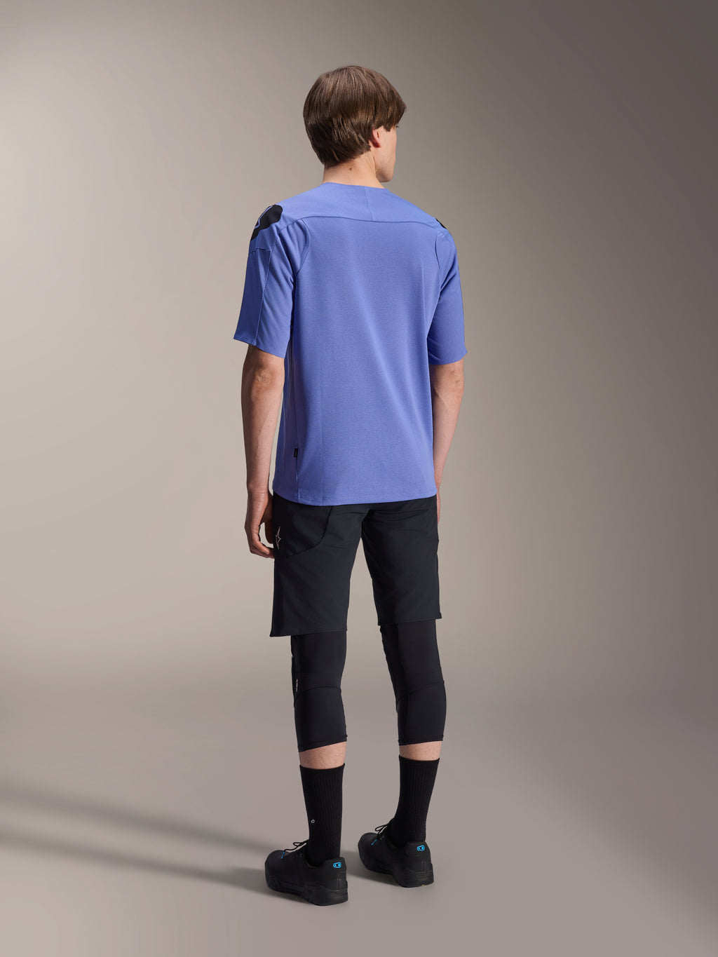 Alpinestars A-Aria Elite Shorts, Mountainbike-Shorts, Black Titanium, Rückansicht getragen mit einem lavendelfarbenen Kurzarmtrikot, schwarzen Knieprotektoren und schwarzen Radschuhen, sportliche MTB-Ausrüstung.