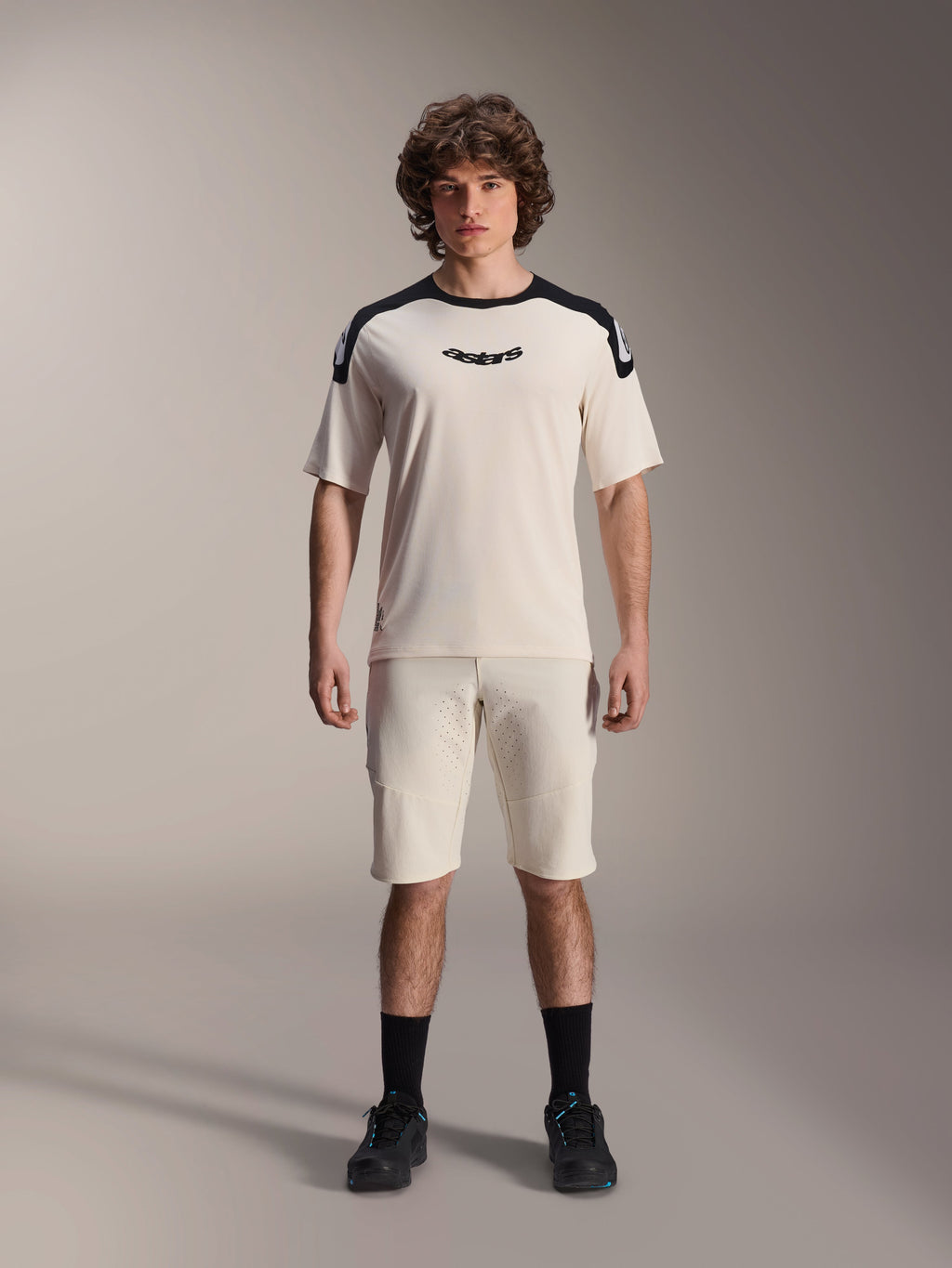 Alpinestars A-Aria Polartec Elite Kurzarmtrikot in Ecru, mit schwarzen Schultereinsätzen und mittigem Brustlogo, kombiniert mit passenden Ecru-Shorts, schwarzen Socken und Radschuhen, hochwertige Mountainbike-Ausrüstung.