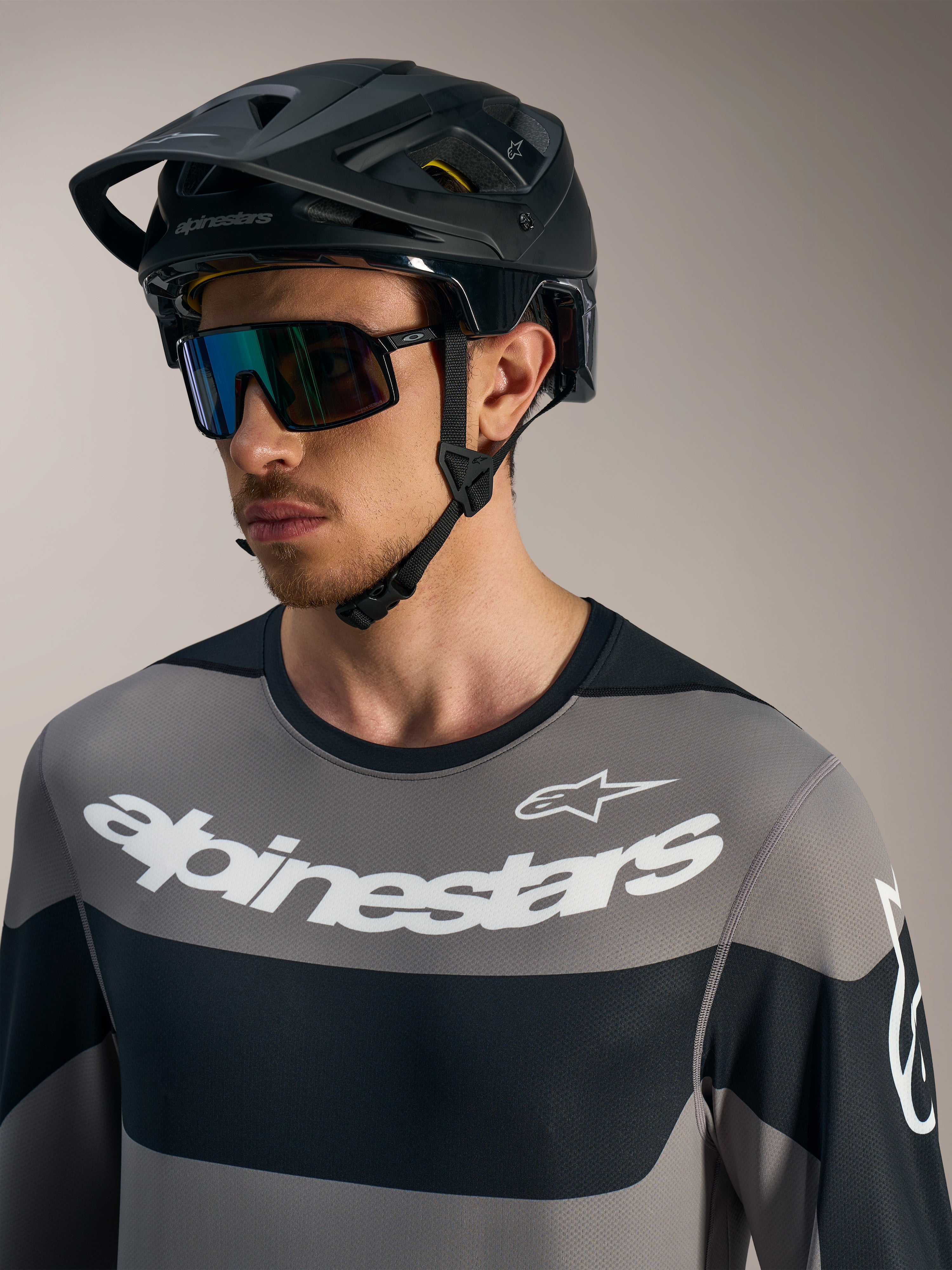 Graues Alpinestars A-Dura Race Langarmtrikot mit weißen Logos, schwarzem MTB-Helm und Performance-Sonnenbrille.