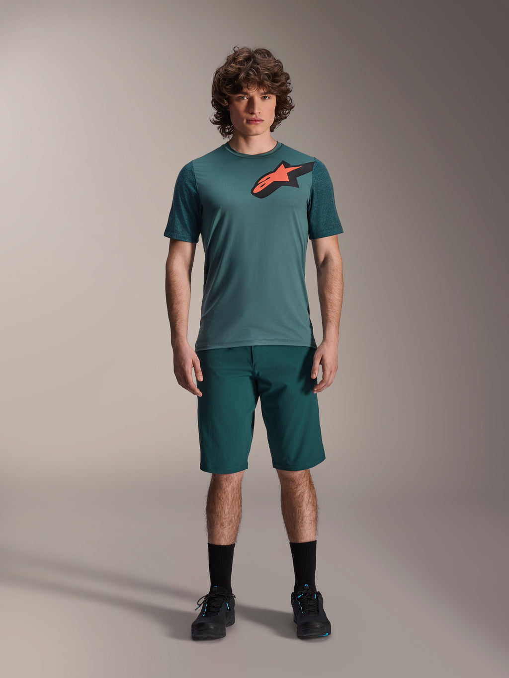 Alpinestars A-Dura Lipan Trikot, Kurzarm-MTB-Trikot, Silver Pine Grün mit schwarz-orangem Astars-Logo auf der Brust, kombiniert mit passenden Shorts und schwarzem Schuhwerk, technische Mountainbike-Bekleidung