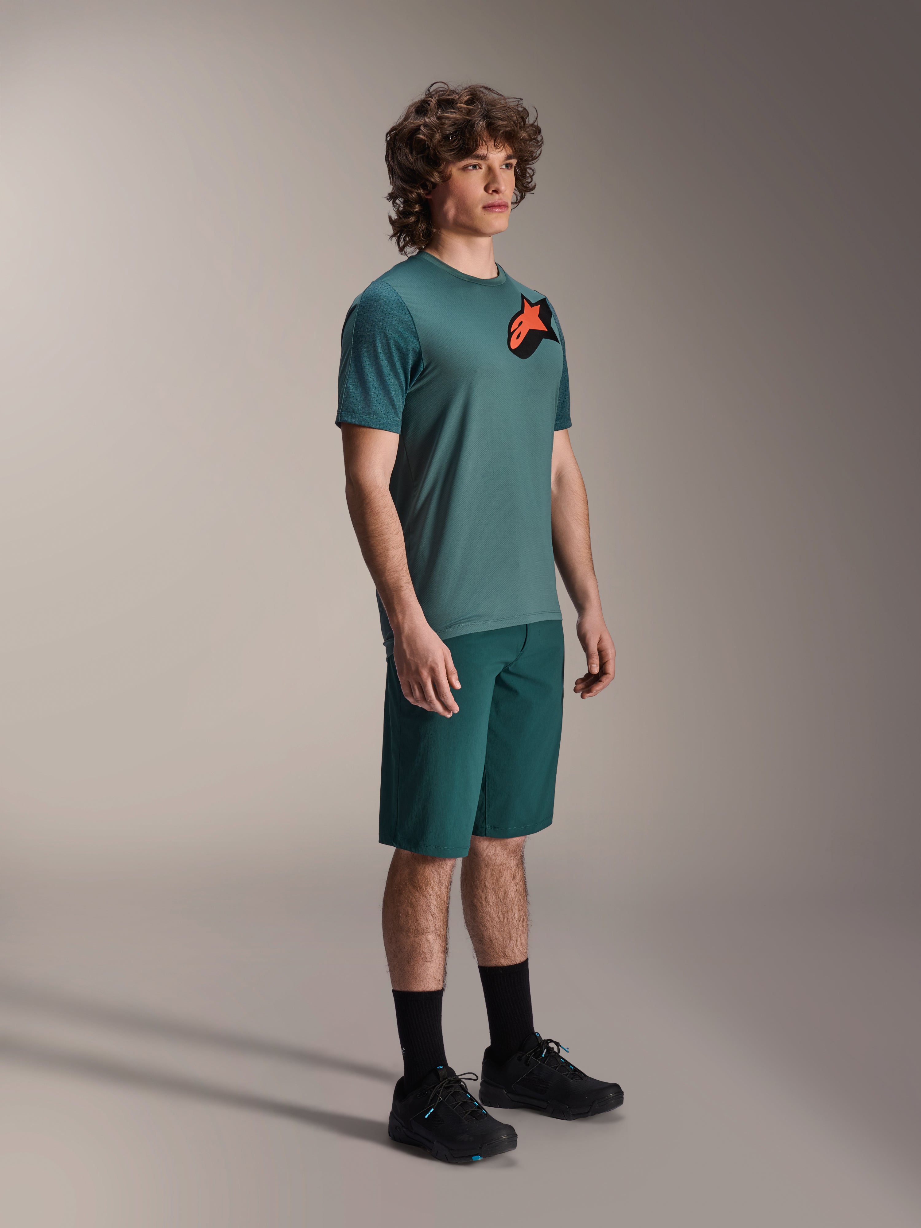 Alpinestars A-Dura Lipan Jersey, kurzärmeliges MTB-Trikot, Silver Pine Grün, mit orange-schwarzem Astars-Logo auf der Brust, kombiniert mit passenden grünen Shorts und schwarzen Radschuhen, lässiger Trail-Stil