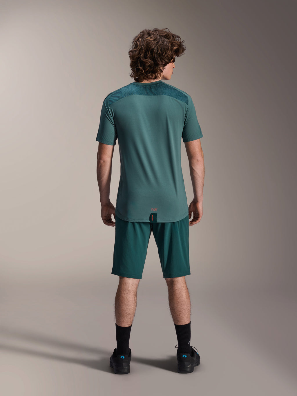 Alpinestars A-Dura Lipan Kurzarmtrikot in Silver Pine, Rückansicht mit texturierten Jacquard-Schulterbahnen und verlängertem Drop-Tail-Saum, getragen mit passenden dunkelgrünen Mountainbike-Shorts und schwarzen Schuhen.