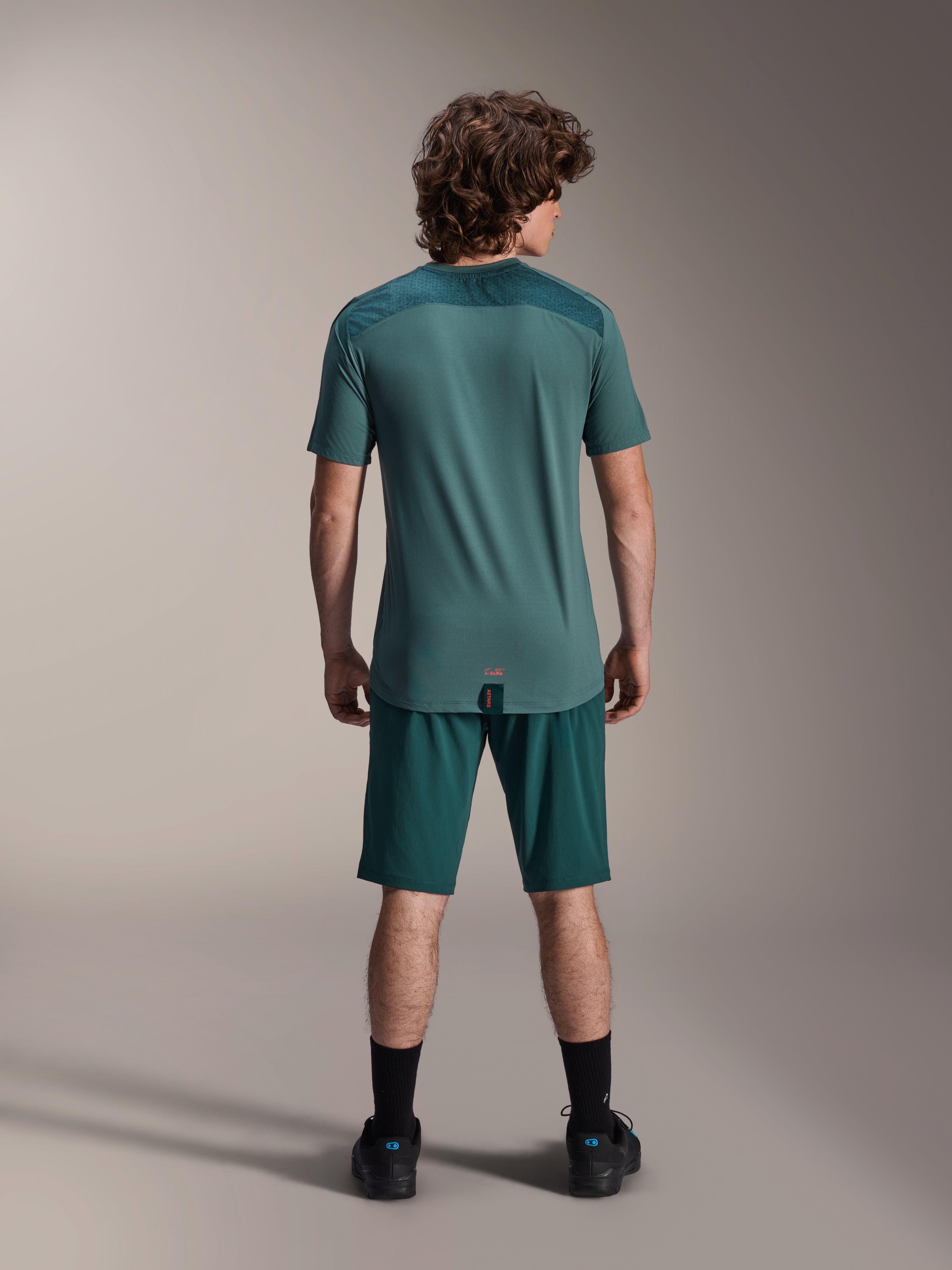 Alpinestars A-Dura Lipan Kurzarmtrikot in Silver Pine, Rückansicht mit texturierten Jacquard-Schulterbahnen und verlängertem Drop-Tail-Saum, getragen mit passenden dunkelgrünen Mountainbike-Shorts und schwarzen Schuhen.