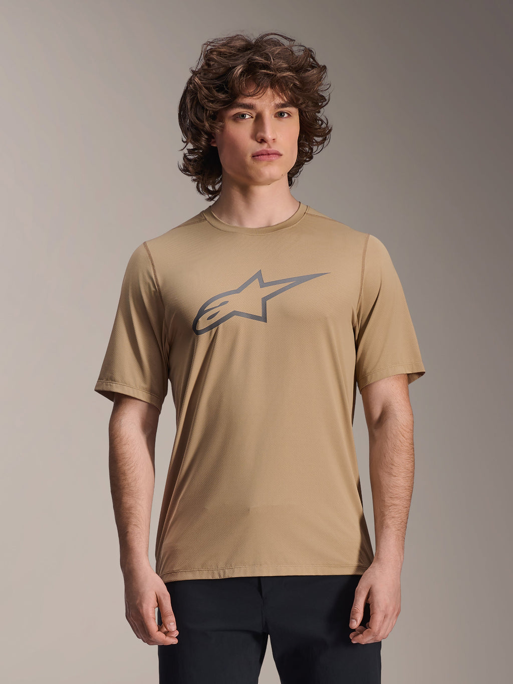 Alpinestars A-Dura Astar Kurzarm-Jersey, Hellbraun, getragen von einem männlichen Model mit schwarzer Hose, graues Alpinestars Stern-Logo auf der Brust, lässiges Mountainbike-Trikot für Trail-Fahrten