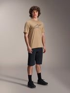 Alpinestars A-Dura Astar Kurzarm-Trikot, Hellbraun, mit einem großen dunkelgrauen Astar-Logo auf der Brust, getragen von einem Modell mit schwarzen MTB-Shorts, schwarzen Socken und schwarzen Schuhen, lässige Performance-Mountainbike-Bekleidung in einer Studio-Umgebung