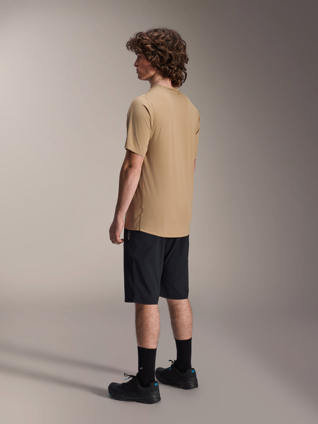 Alpinestars A-Dura Liner Shorts in Schwarz und A-Dura SS Jersey in Tan, getragen von einem männlichen Model in der Seitenprofilansicht, kombiniert mit schwarzen Socken und Radschuhen, Mountainbike-Lifestyle-Bekleidung.