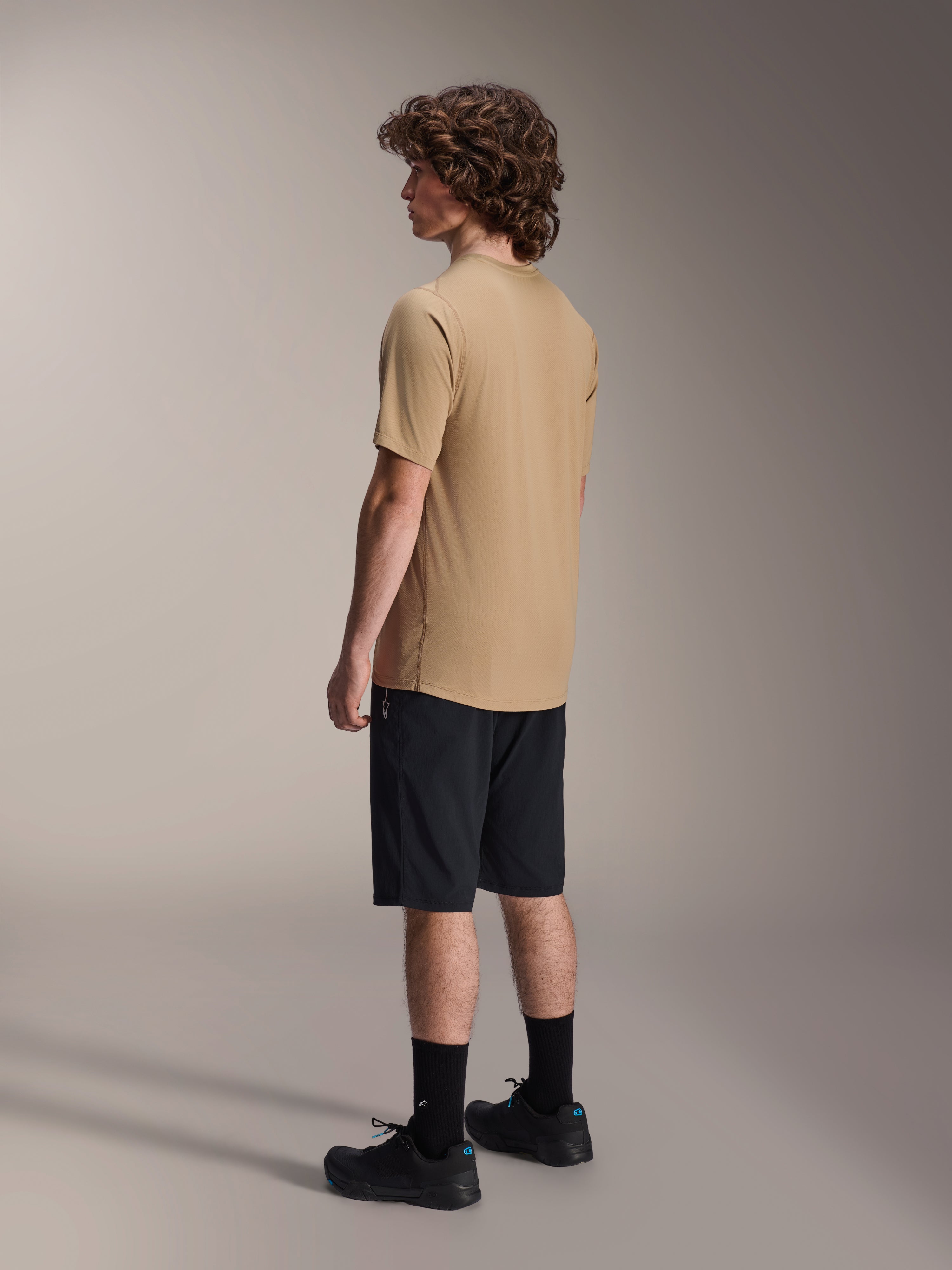 Alpinestars A-Dura Liner Shorts in Schwarz und A-Dura SS Jersey in Tan, getragen von einem männlichen Model in der Seitenprofilansicht, kombiniert mit schwarzen Socken und Radschuhen, Mountainbike-Lifestyle-Bekleidung.