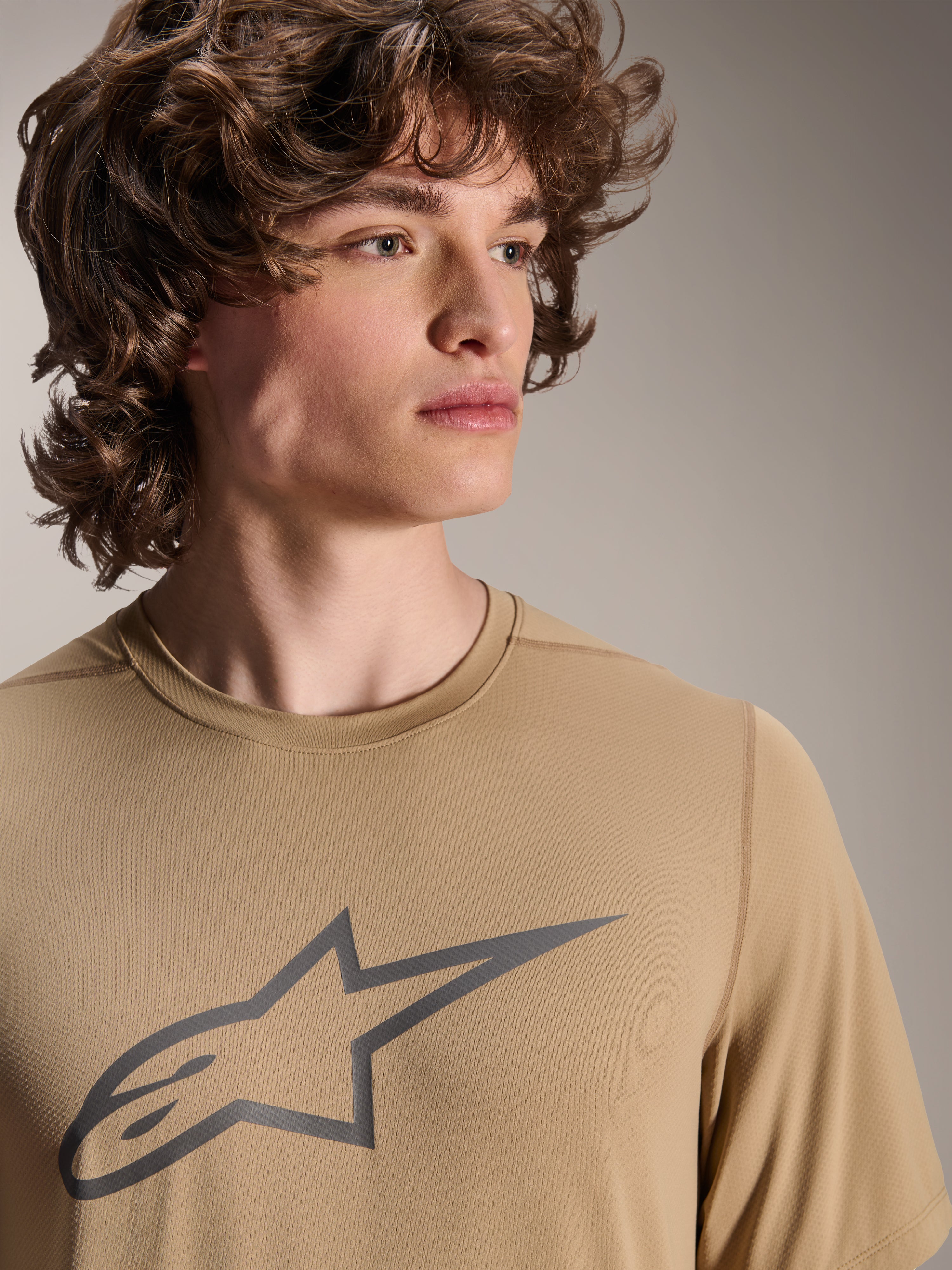 Modell trägt Alpinestars A-Dura Astar Kurzarm-Trikot, hellbraunes MTB-Trikot, mit grauem Stern-Logo auf der Brust, feuchtigkeitsableitendes Jacquard-Gewebe, lässiger Radsport-Stil