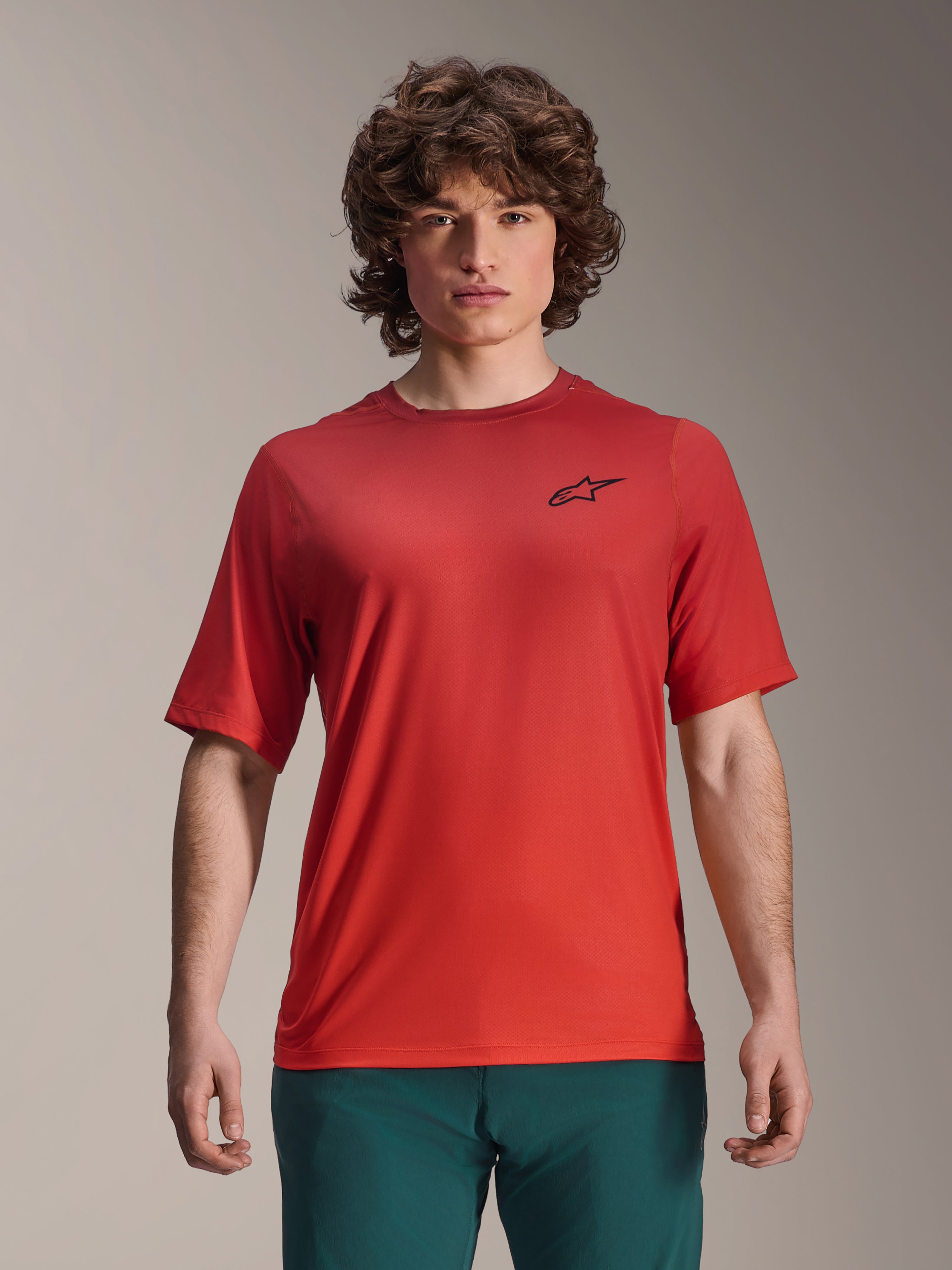 Alpinestars 2025 A-Dura Rise Trikot, Kurzarm-MTB-Shirt, Spicy Orange, von einem Mann getragen, mit schwarzem Astars-Logo auf der Brust, kombiniert mit dunkelgrüner Hose, lässige Performance-Mountainbike-Bekleidung