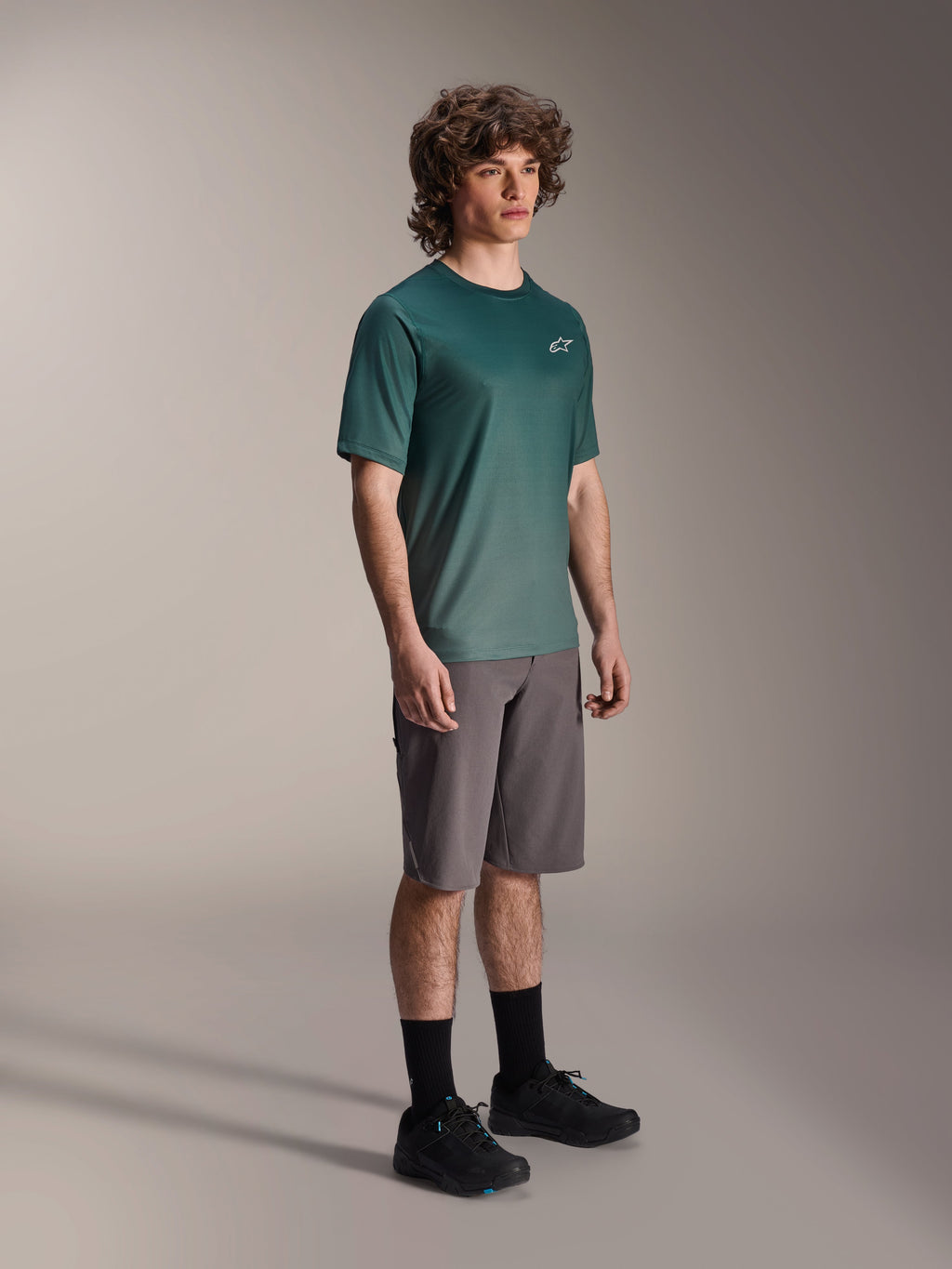 Alpinestars A-Dura Rise Kurzarmtrikot in Silver Pine, grünes Mountainbike-Trikot getragen von einem Model mit grauen Shorts und schwarzen Sneakern, lässige MTB-Trail-Ausrüstung mit kleinem weißen Logo auf der Brust