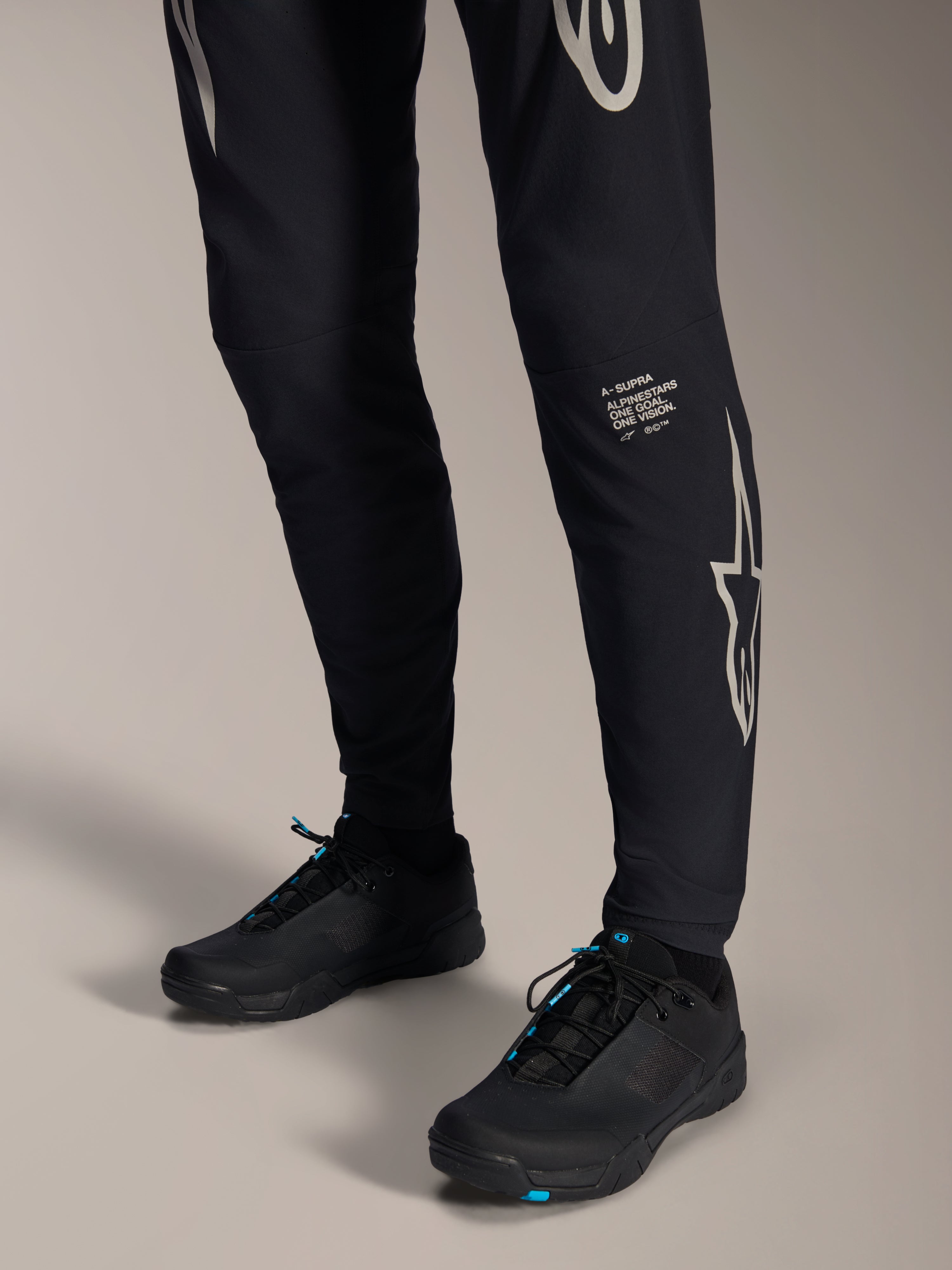 Alpinestars A-Supra Melt Hose, schwarze Mountainbike-Hose, weißer Alpinestars-Schriftzug und Logo auf dem Bein, schwarze Fahrradschuhe mit blauen Akzenten, Performance-Offroad-Ausrüstung