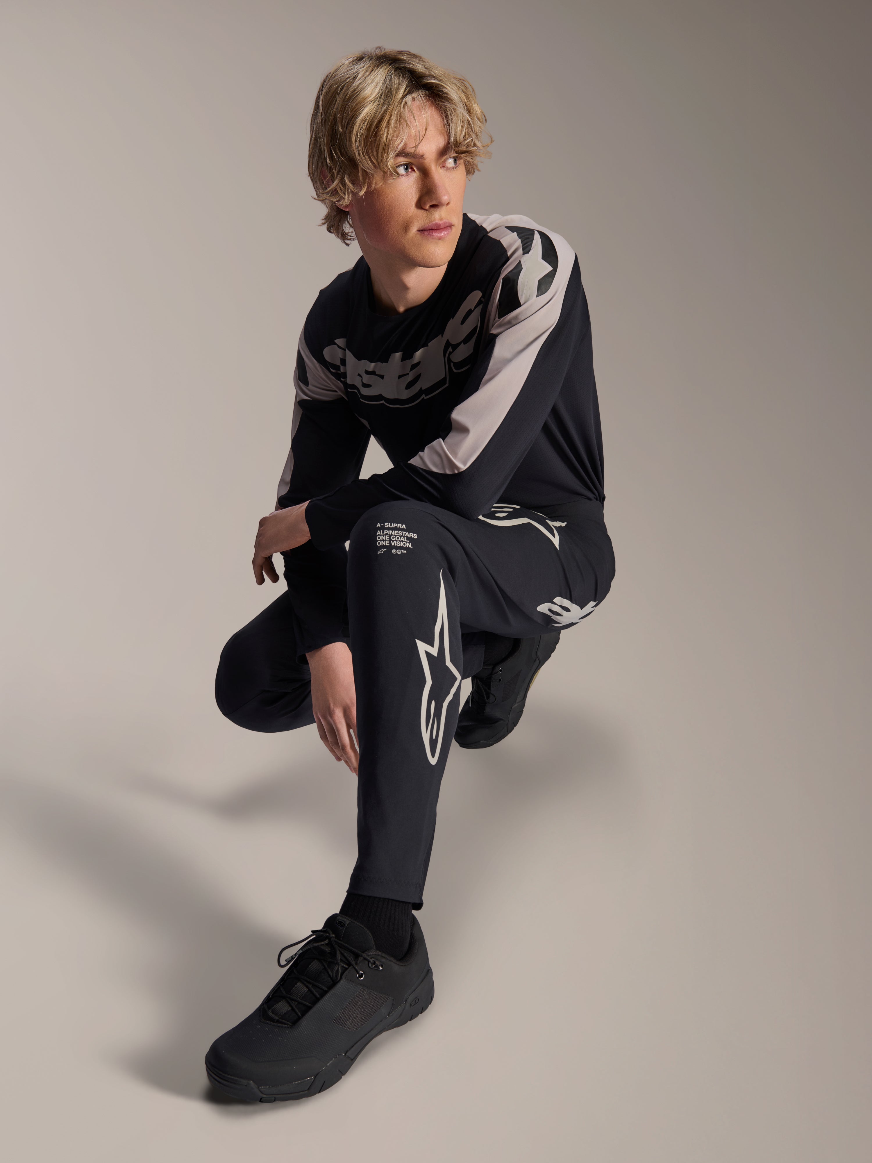 Alpinestars A-Supra Melt Hose und Langarmtrikot, Schwarz und Grau, mit auffälligem weißem Branding und Stern-Logos, getragen von einem männlichen Model in hockender Pose, abgestimmte MTB-Rennausrüstung