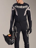 Alpinestars A-Supra Melt Langarm-Trikot, Schwarz mit grauen Grafik-Panels und markantem „astars“-Schriftzug auf der Brust, getragen mit schwarzer Mountainbike-Hose und Handschuhen, hält einen schwarzen Full-Face-Helm, Performance-MTB-Rennausrüstung