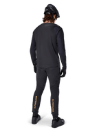 Rear view of a mountain bike rider wearing the Alpinestars A-Aria Polartec Switch Long Sleeve Jersey in Black, paired with black MTB pants featuring tan Alpinestars branding on the calves, black helmet and gloves, professional cycling gear for off-road trails.