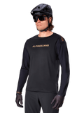 Alpinestars A-Aria Polartec Switch Long Sleeve Jersey, Black with gold logo, MTB mountain biking apparel, worn with a black helmet and sunglasses, featuring laser-cut ventilation on the sleeves and moisture-wicking Polartec fabric for off-road performance