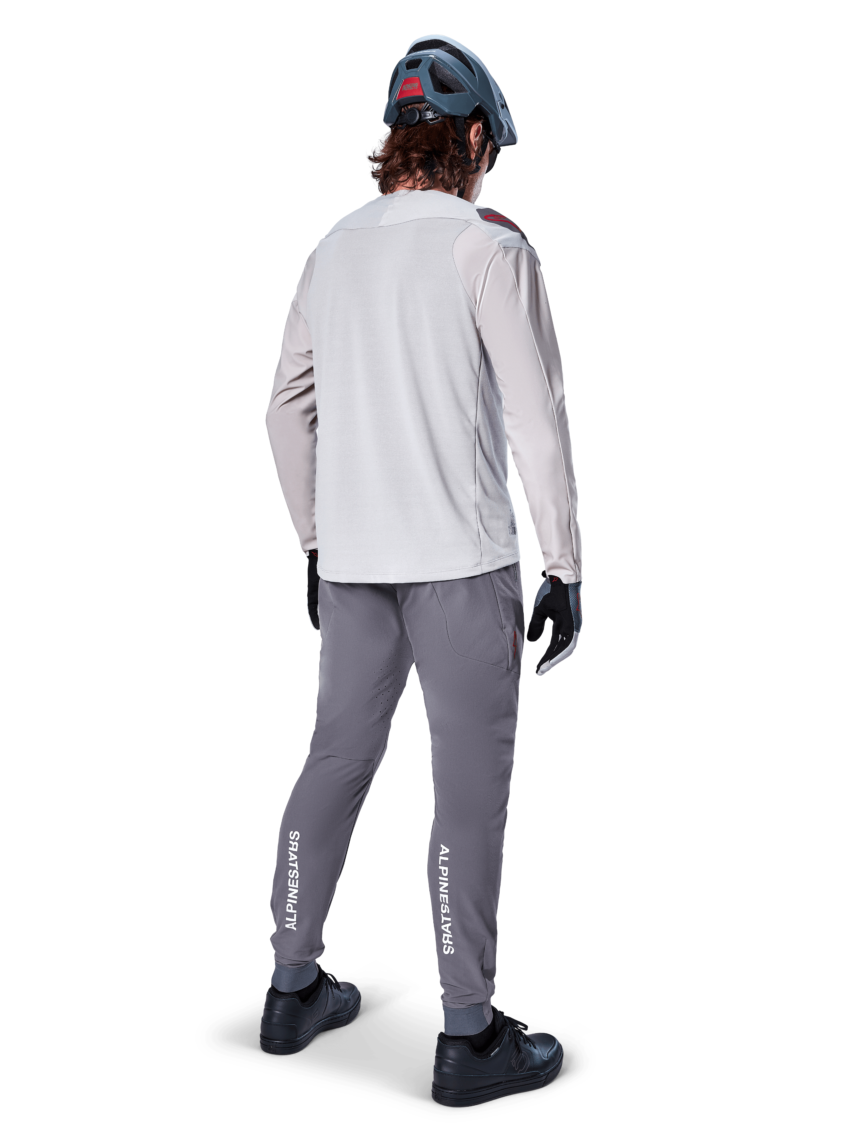 Rear view of a cyclist wearing the Alpinestars A-Aria Polartec Switch Long Sleeve Jersey in Light Gray, paired with dark gray mountain bike pants featuring white Alpinestars branding on the calves, black gloves, and a gray helmet.