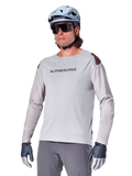 Alpinestars A-Aria Polartec Switch Jersey Long Sleeve, Light Gray cycling jersey with black chest logo and red shoulder accent, laser-perforated sleeves for airflow, worn with a light blue helmet, sunglasses, and gray pants, performance mountain bike apparel for trail riding
