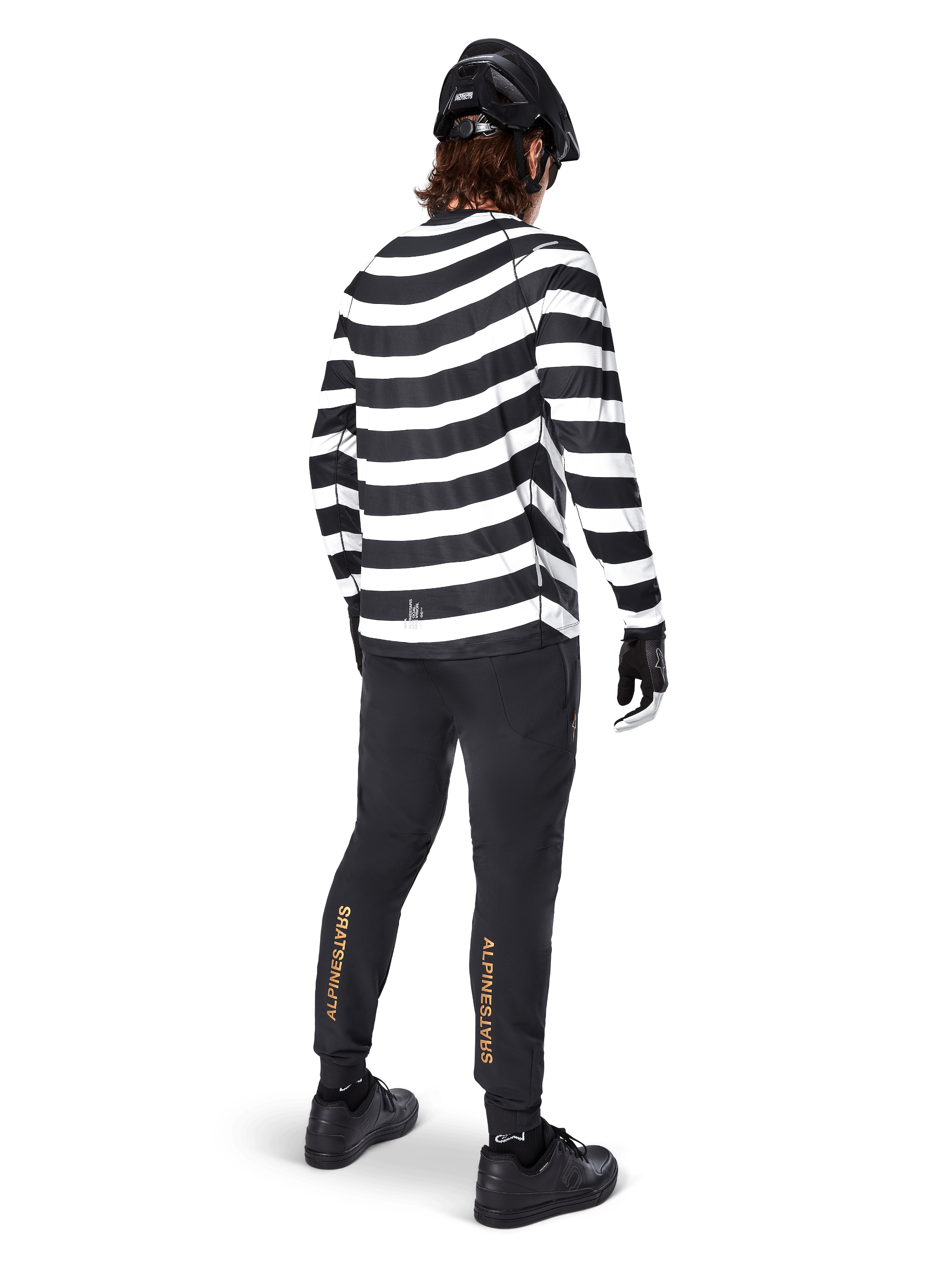 MTB rider wearing the Alpinestars A-Aria Oscar Long Sleeve Jersey, black and white horizontal spiral stripes, paired with black technical pants featuring gold logos on the calves, black helmet and shoes, complete off-road cycling outfit from behind