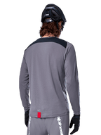 Alpinestars A-Dura Dri Astar Langarmtrikot, dunkelgraues MTB-Trikot von hinten gezeigt, mit schwarzem Schulterbereich und kleinem roten Markenlabel, kombiniert mit schwarzem Mountainbike-Helm und Handschuhen.